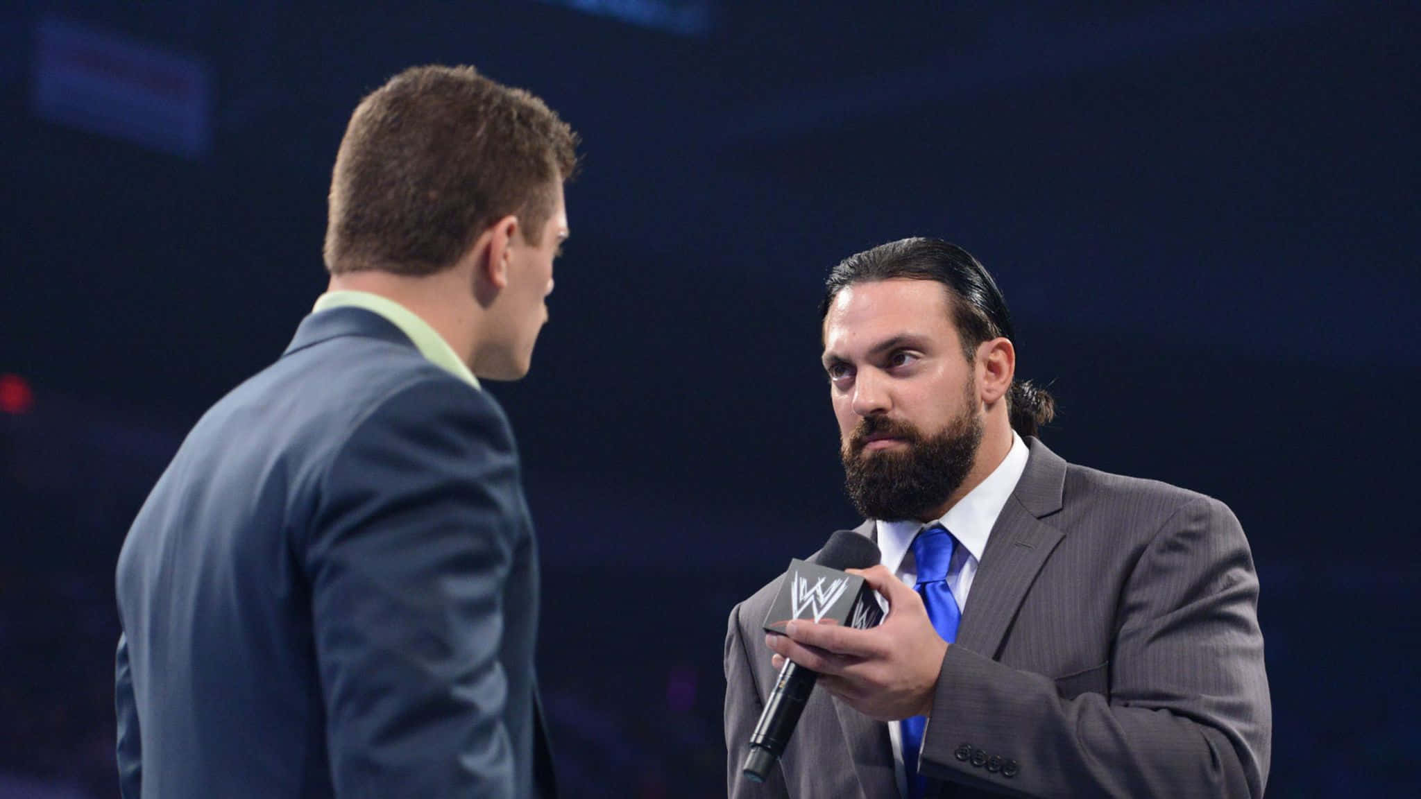 WWE 桌布 壁紙 — WWE摔角手留鬍鬚穿西裝，在燈光昏暗的場館內面向採訪者手持麥克風。