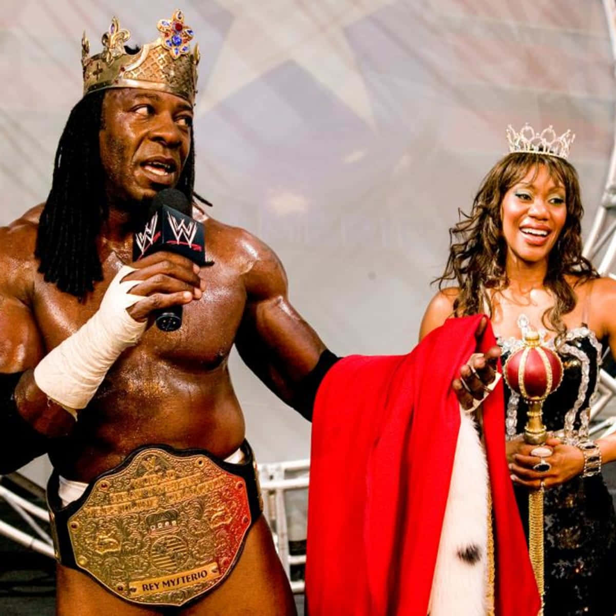 WWE 桌布 壁紙 — WWE摔角手雷伊·米斯特里奧戴著王冠和冠軍腰帶，站在一位戴著頭飾、微笑的女子旁邊，女子手持權杖和紅色斗篷。