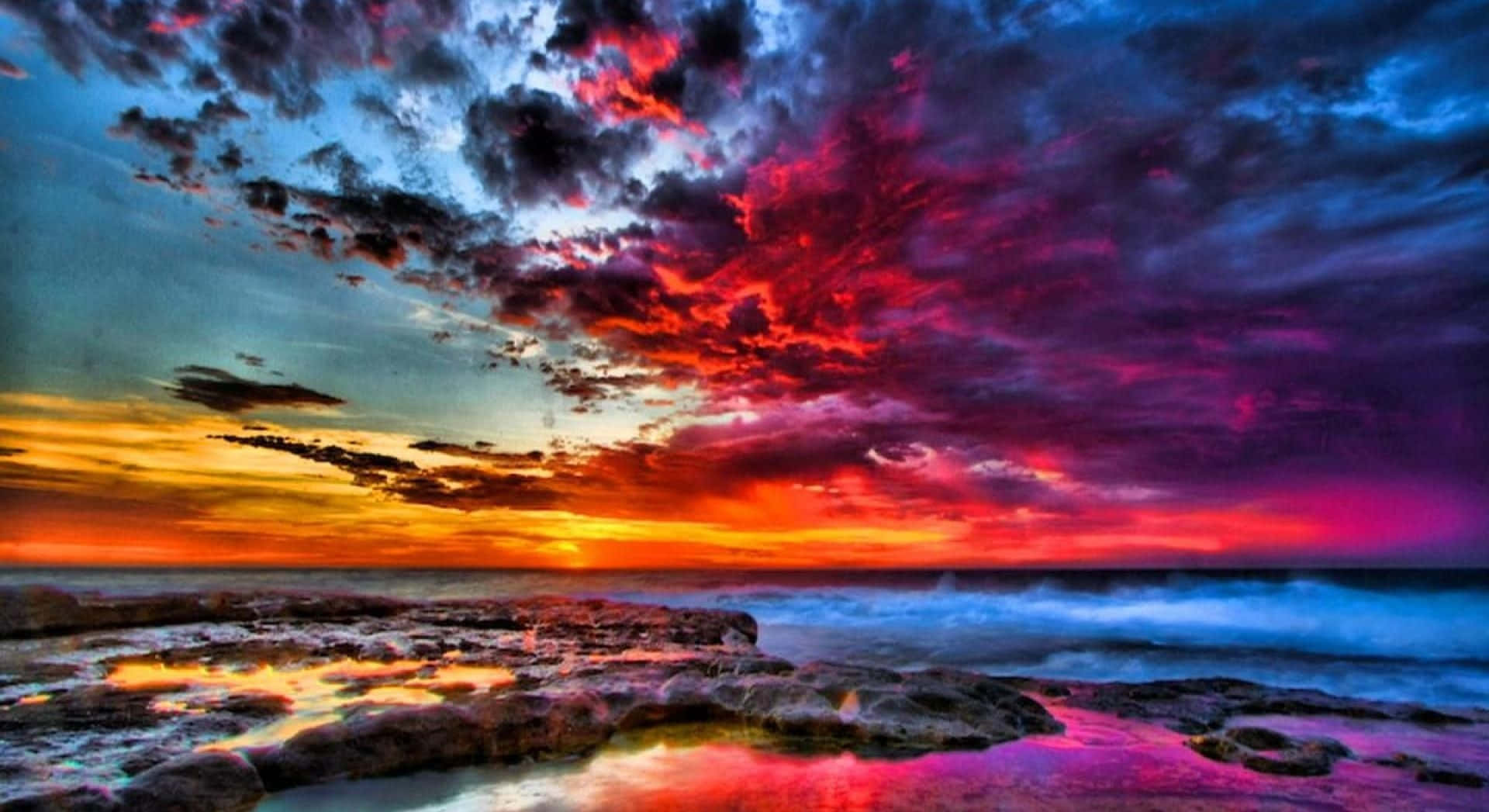 Colorful Sunset 桌布 壁紙 — 戲劇性的日落將天空染成火紅、紫色和橙色，映照在岩石海岸線和拍岸的波浪之上。