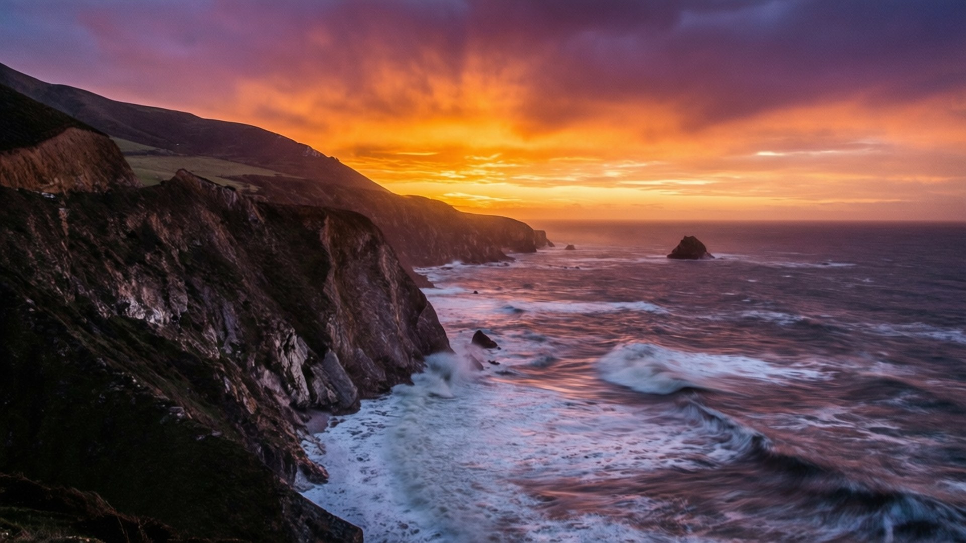 Jurassic Coast Sunset 壁紙 — 戲劇性的日落將天空染成火熱的橙色和紫色，海浪拍打著俯瞰海洋的崎嶇懸崖。