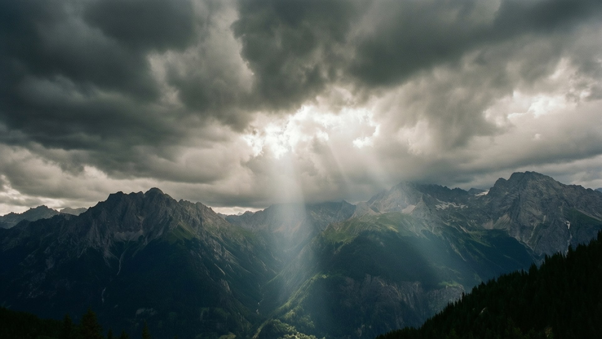 Mountains Under Storm 壁紙 — 戲劇性的山峰刺穿黑暗的暴風雲，燦爛的陽光如瀑布般灑落在森林覆蓋的山坡上。