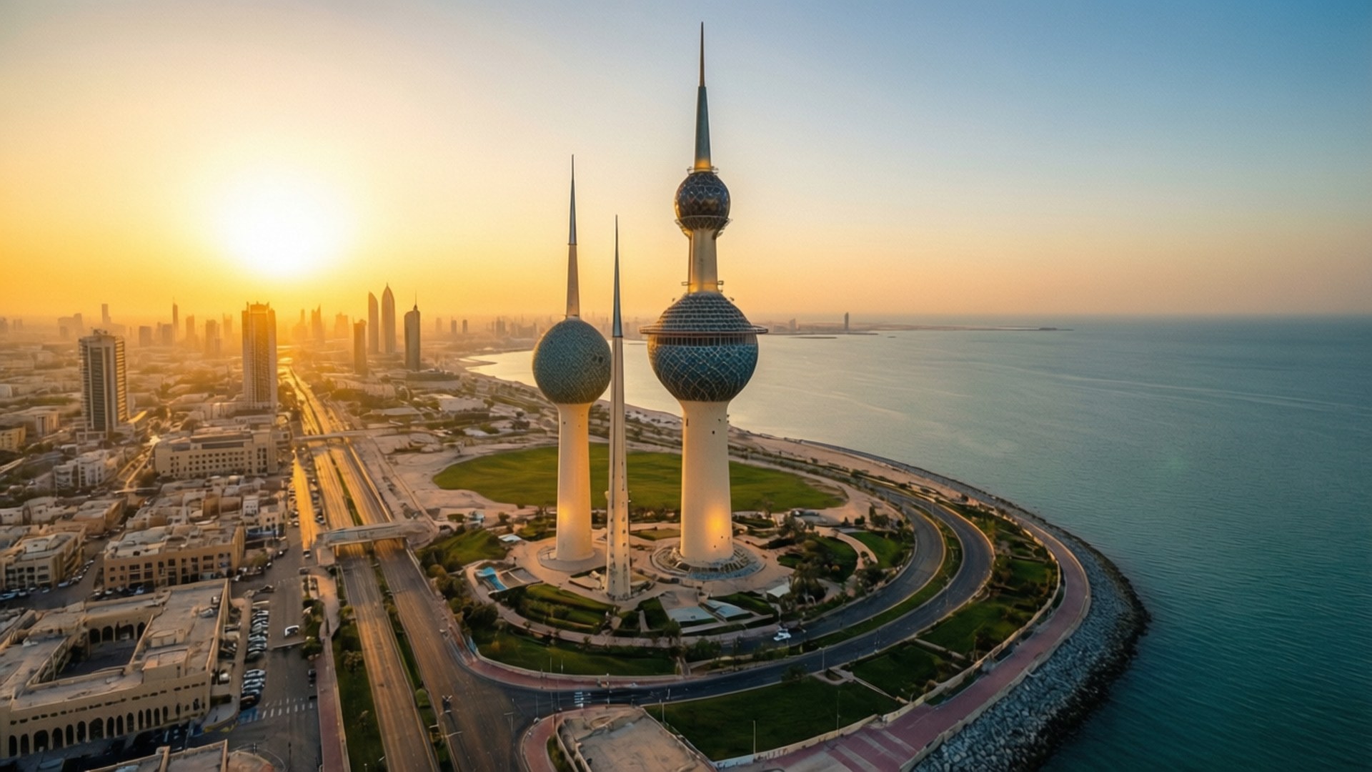Kuwait City Sunrise Aerial 壁紙 — 夕陽將科威特塔沐浴在金黃色光芒中，它們在平靜的藍色海水旁的海岸城市景觀上空升起。
