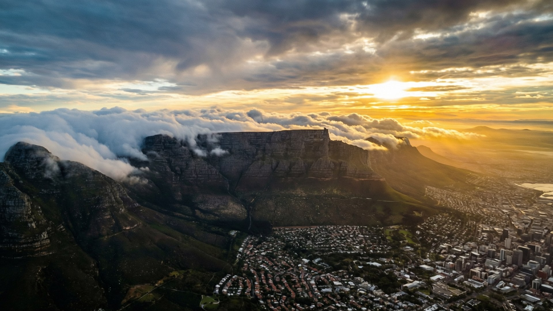 Table Mountain Golden Dawn 壁紙 — 夕陽將桌山沐浴在金色光芒中，雲朵在其平坦的山頂下方旋轉，俯瞰著一座廣闊的城市。