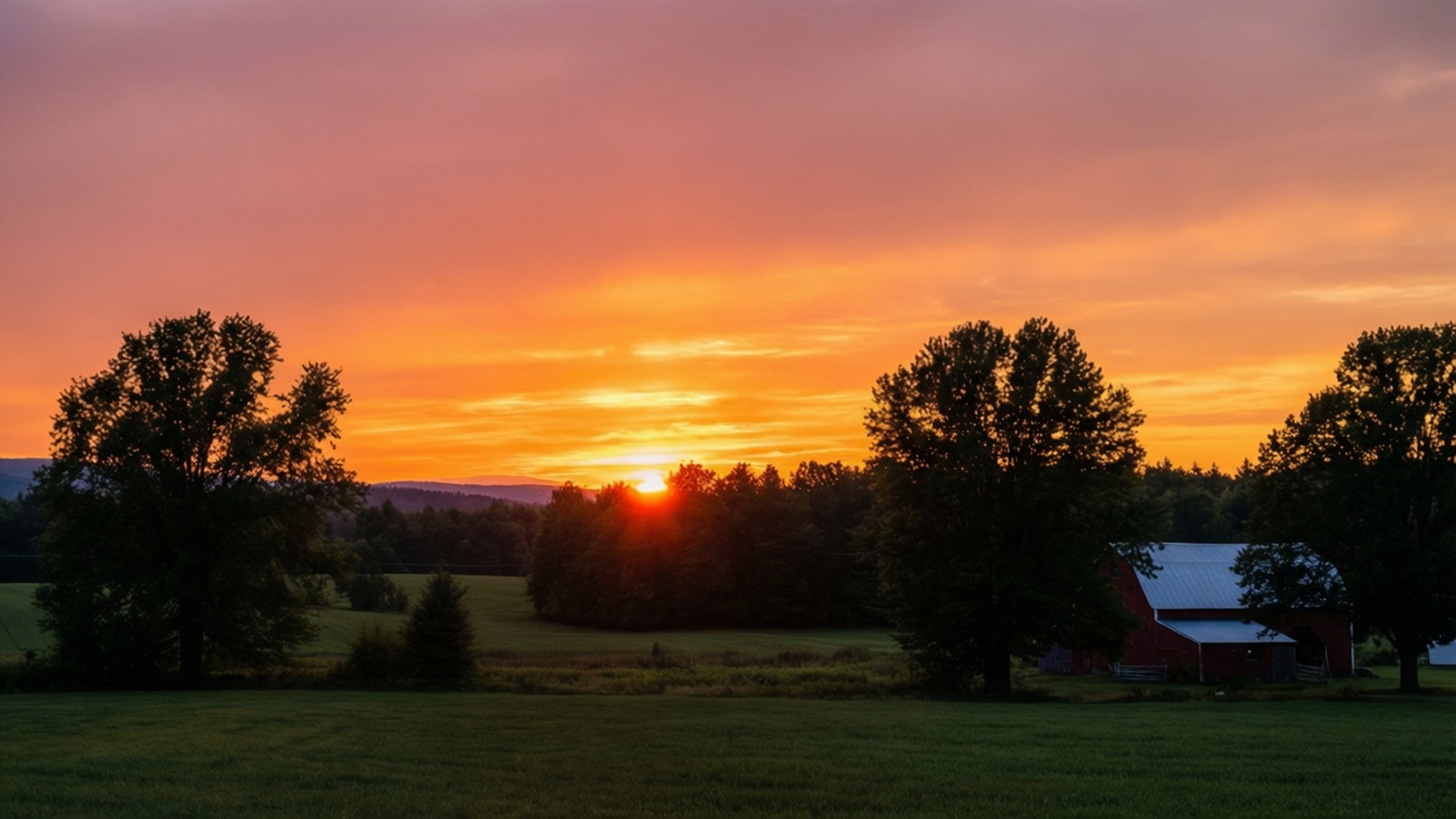 Vermont Rural Sunset 壁紙 — 夕陽西下，將天空染成火熱的橙色和粉紅色，落在紅穀倉和綠色田野襯托下的一排樹木剪影之後。