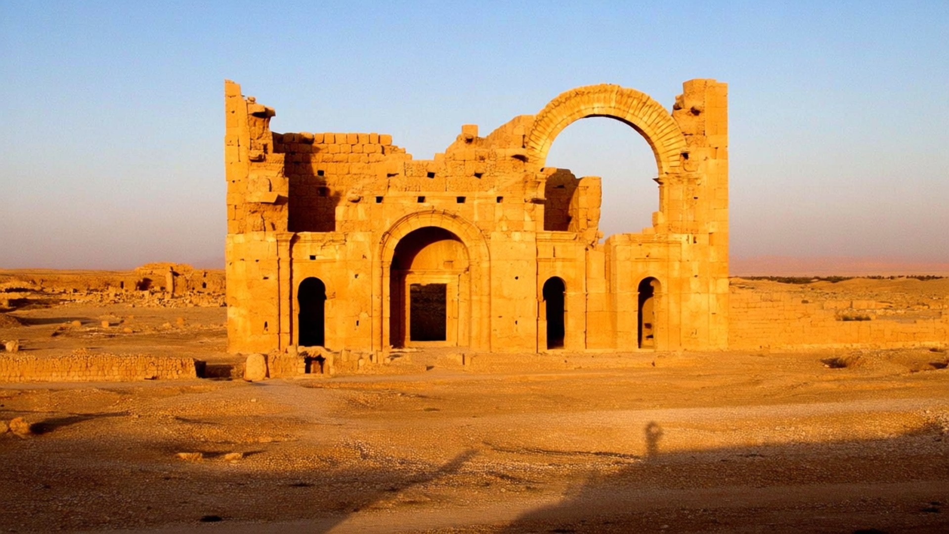 Hatra Ruins Golden Hour 壁紙 — 夕陽以金色光芒沐浴著古老的石頭廢墟，陰影在沙漠地面上延伸。