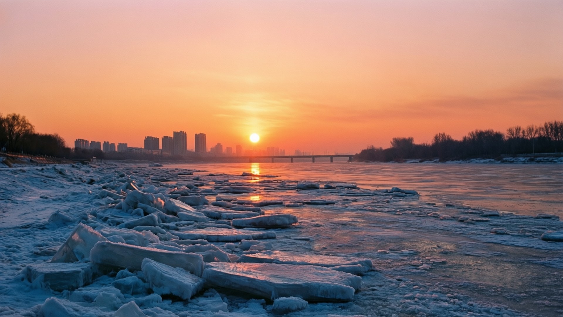 River Sunrise 壁紙 — 夕陽在結冰的河上投射出溫暖的橙色光芒，浮冰和遠處的城市天際線。