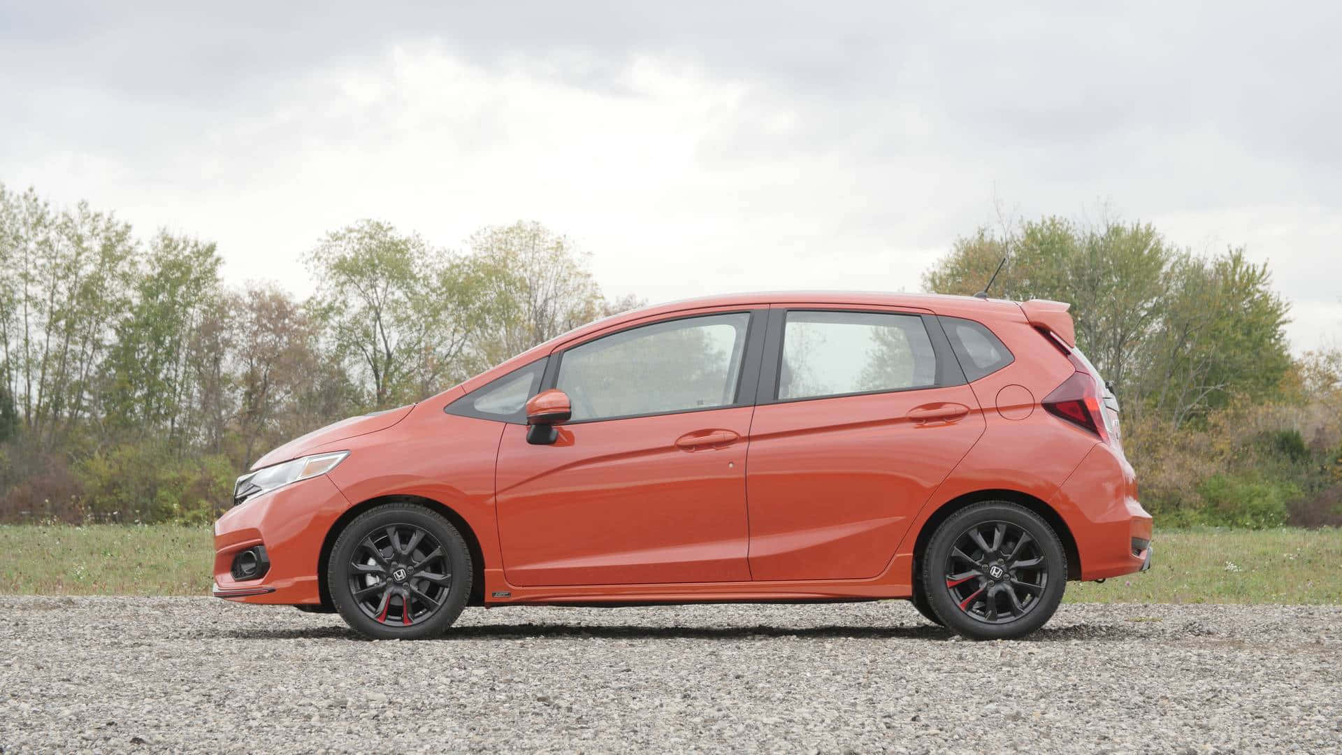 Honda Fit 桌布 壁紙 — 一輛鮮橙色的本田Fit搭配黑色輪圈，停放在碎石上，旁邊有一排樹木，天空多雲。