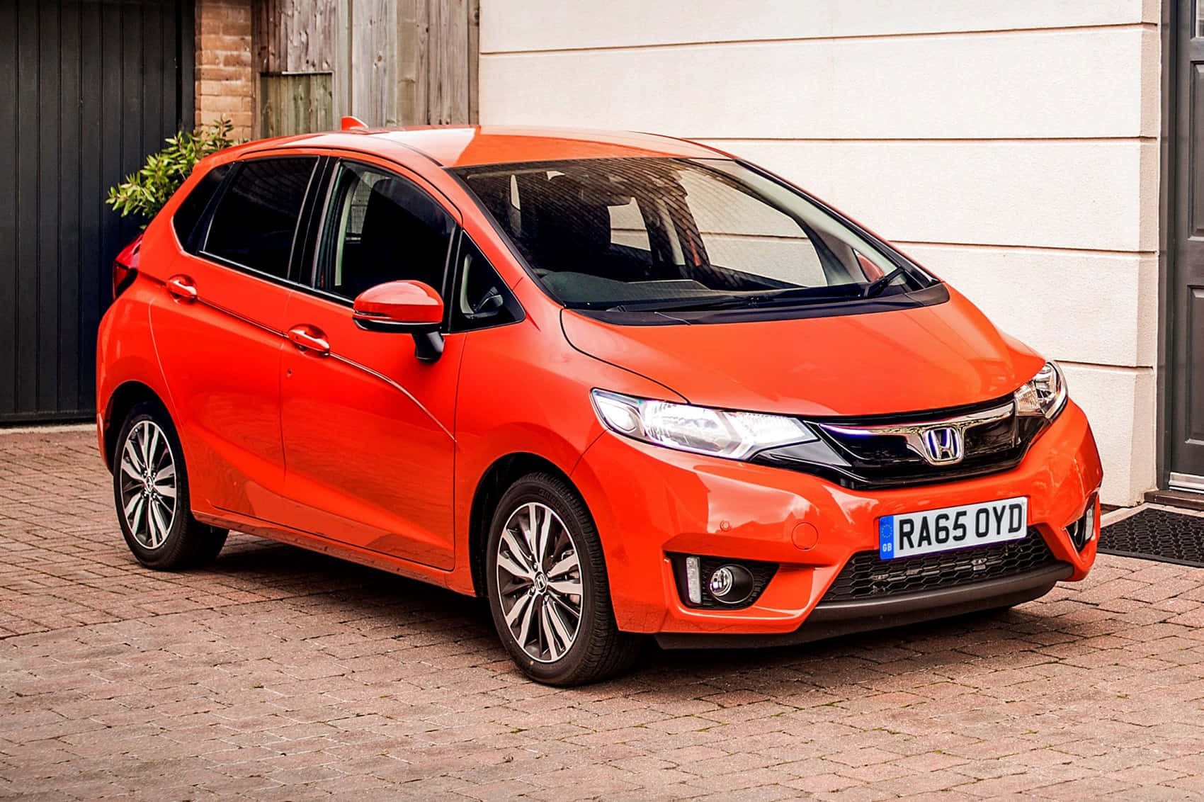 Honda Fit 桌布 壁紙 — 一輛鮮橙色的本田 Jazz 停放在磚砌車道上，旁邊是一棟帶木質裝飾的現代房屋。