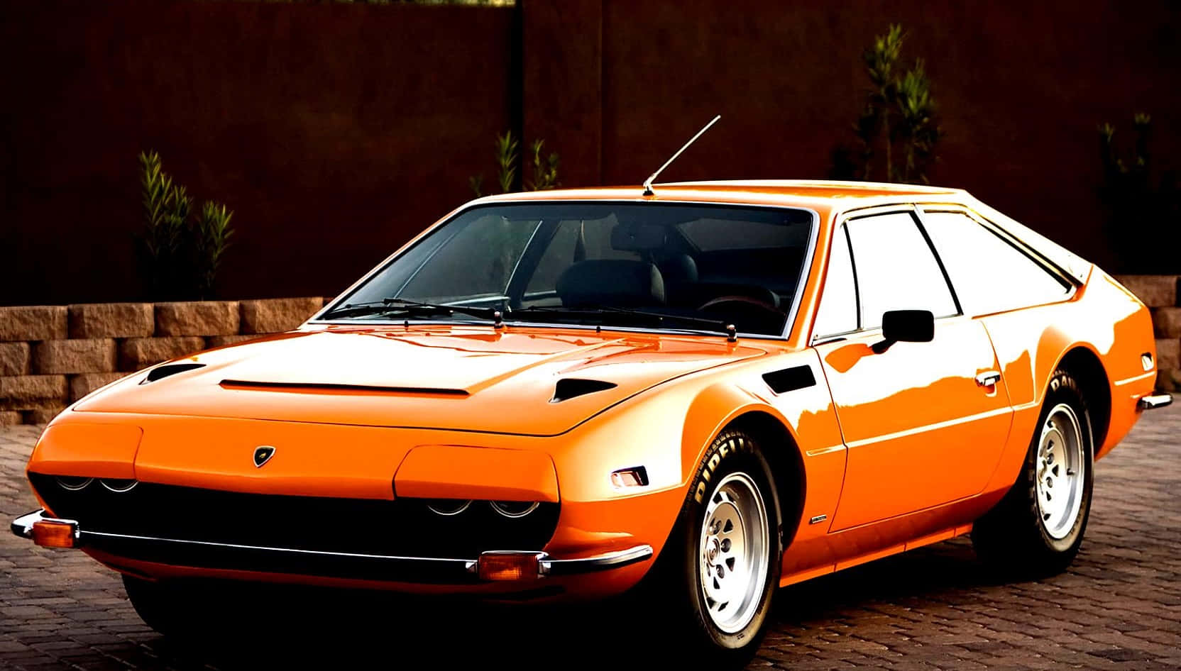 Lamborghini Silhouette 桌布 壁紙 — 一輛鮮橙色的經典藍寶堅尼Countach停放在磚砌車道上，背景有石牆和綠植。