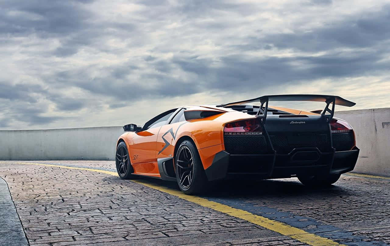 Lamborghini Murciélago 桌布 壁紙 — 一輛鮮橙色的藍寶堅尼 Murciélago SV 停泊在鵝卵石路上，天空烏雲密佈，氣氛戲劇化。