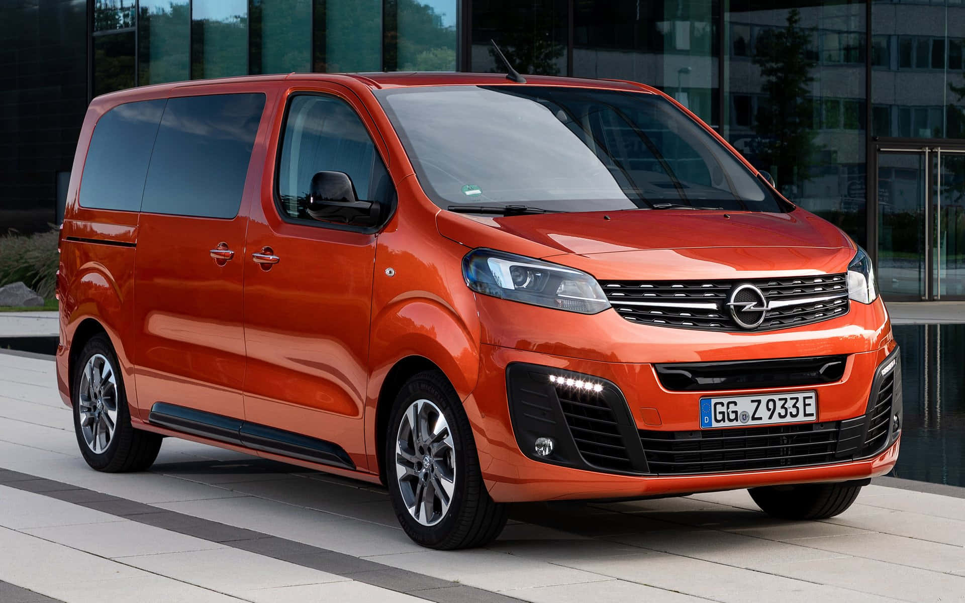 Opel Zafira 桌布 壁紙 — 一輛鮮橙色的歐寶Zafira Tourer時尚地停放在現代磁磚廣場上，旁邊是一座玻璃建築。