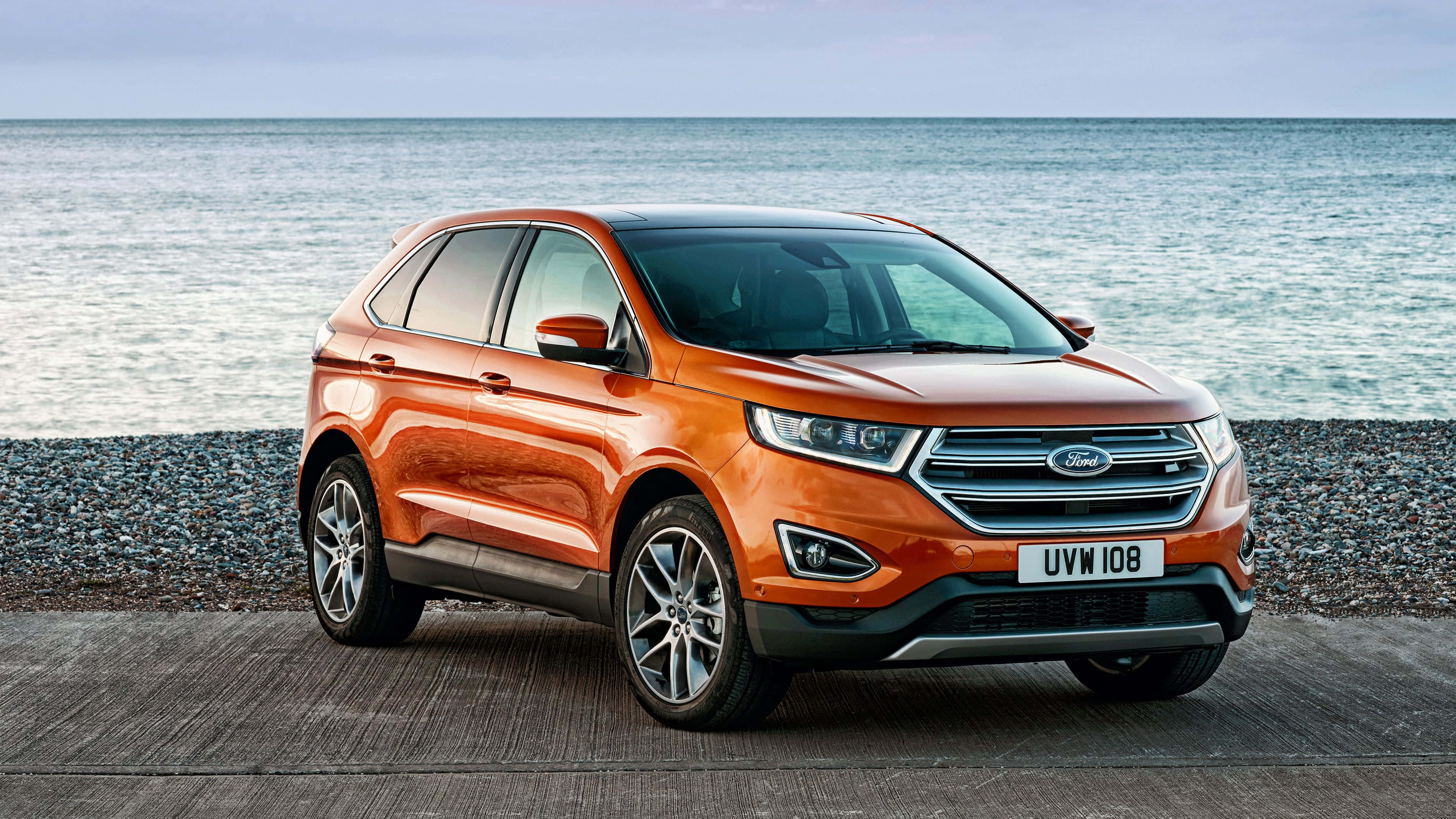 Ford Edge 桌布 壁紙 — 一輛鮮橙色的福特Edge SUV停泊在木製碼頭上，卵石海灘和寧靜的海洋在陰天延伸至地平線。
