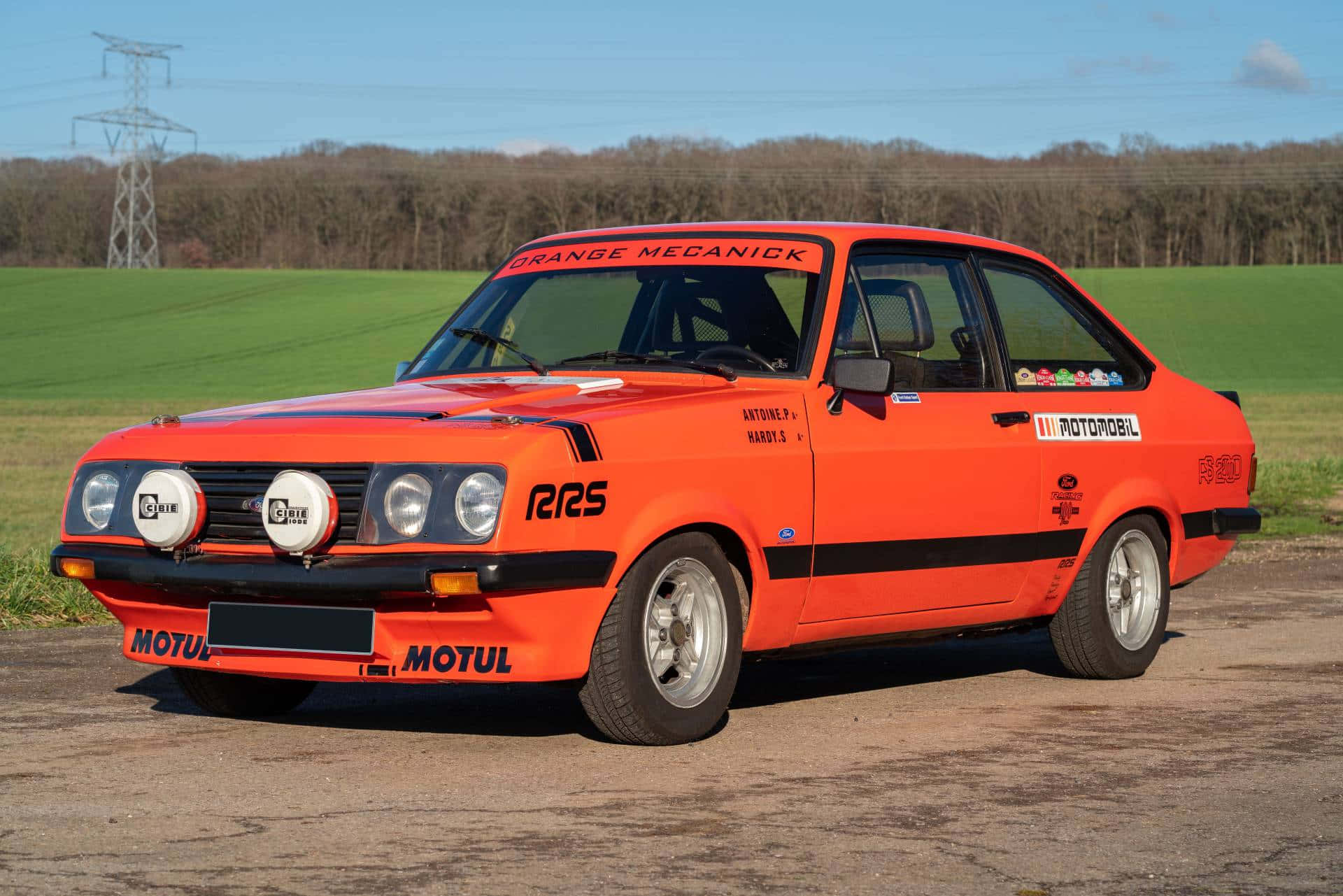 Ford Escort 桌布 壁紙 — 一輛鮮橙色的經典拉力賽車，帶有黑色賽車條紋和贊助商貼紙，停在鄉村田野的碎石上，天空湛藍。