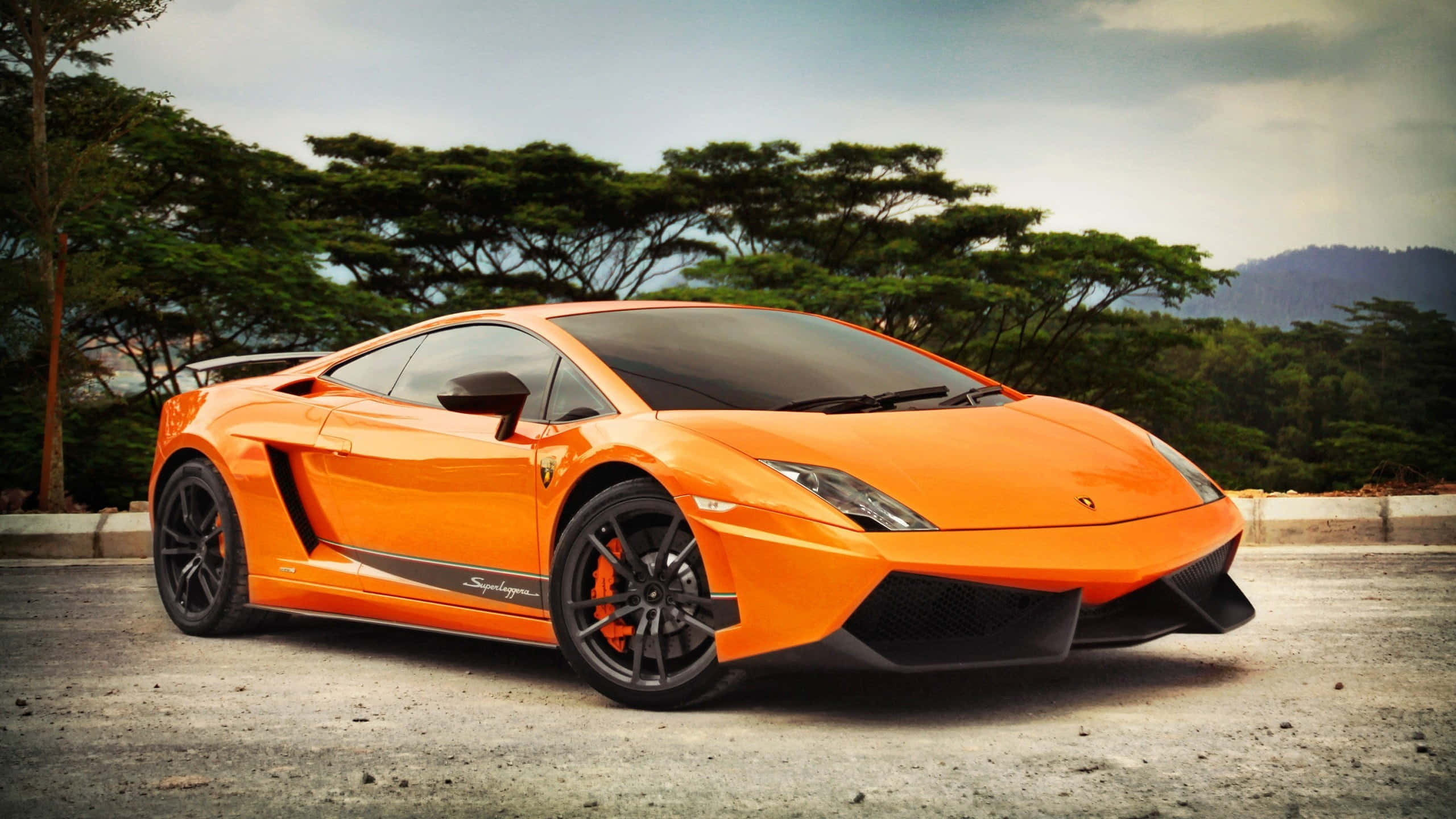 Lamborghini Gallardo 桌布 壁紙 — 一輛鮮橙色藍寶堅尼Gallardo Superleggera停泊在碎石路上，周圍有茂密的綠樹和遠山，天空多雲。
