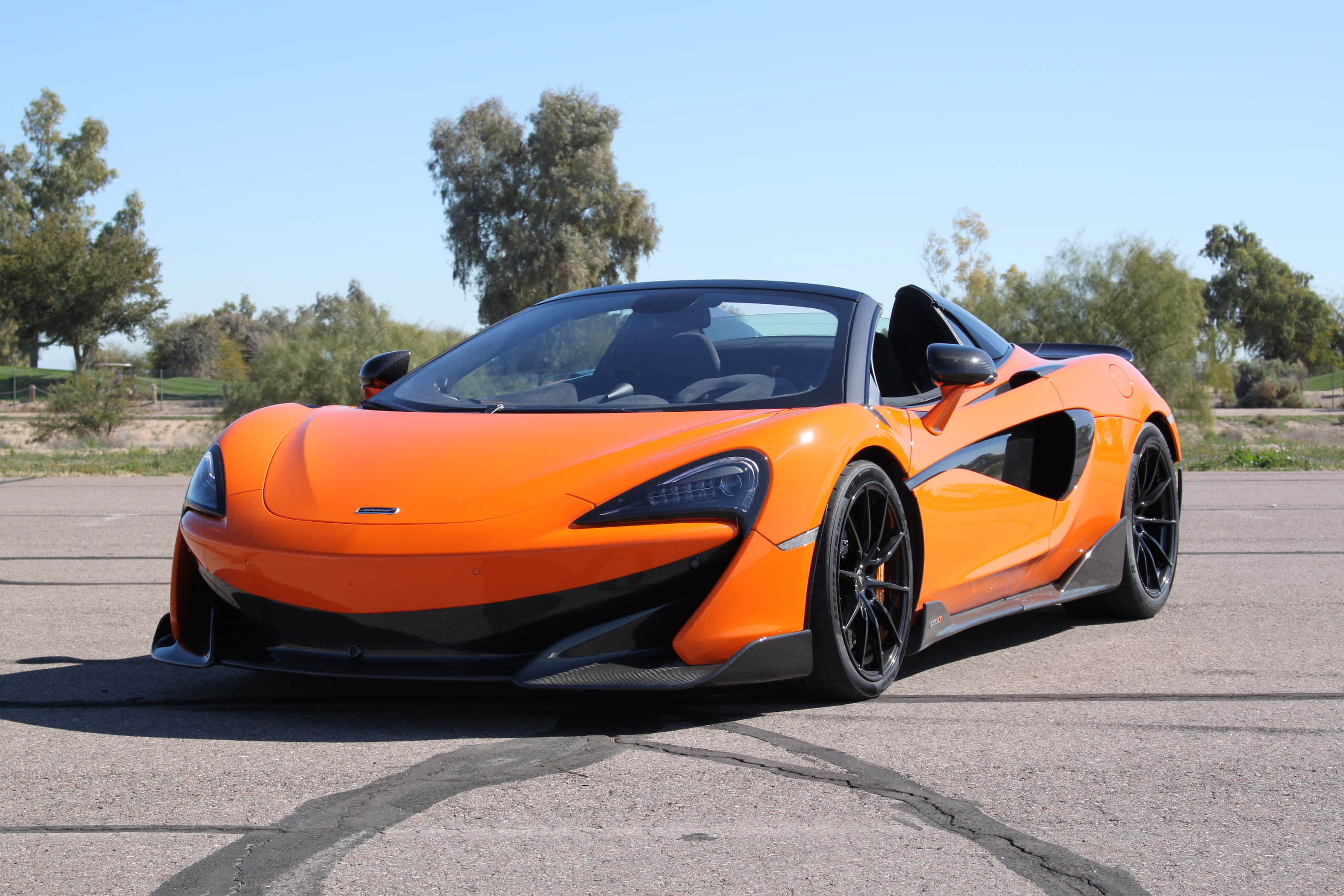Mclaren 600lt Spider 桌布 壁紙 — 一輛鮮橙色的McLaren 570S Spider停放在裂開的柏油路上，背景是清澈的藍天和樹木。