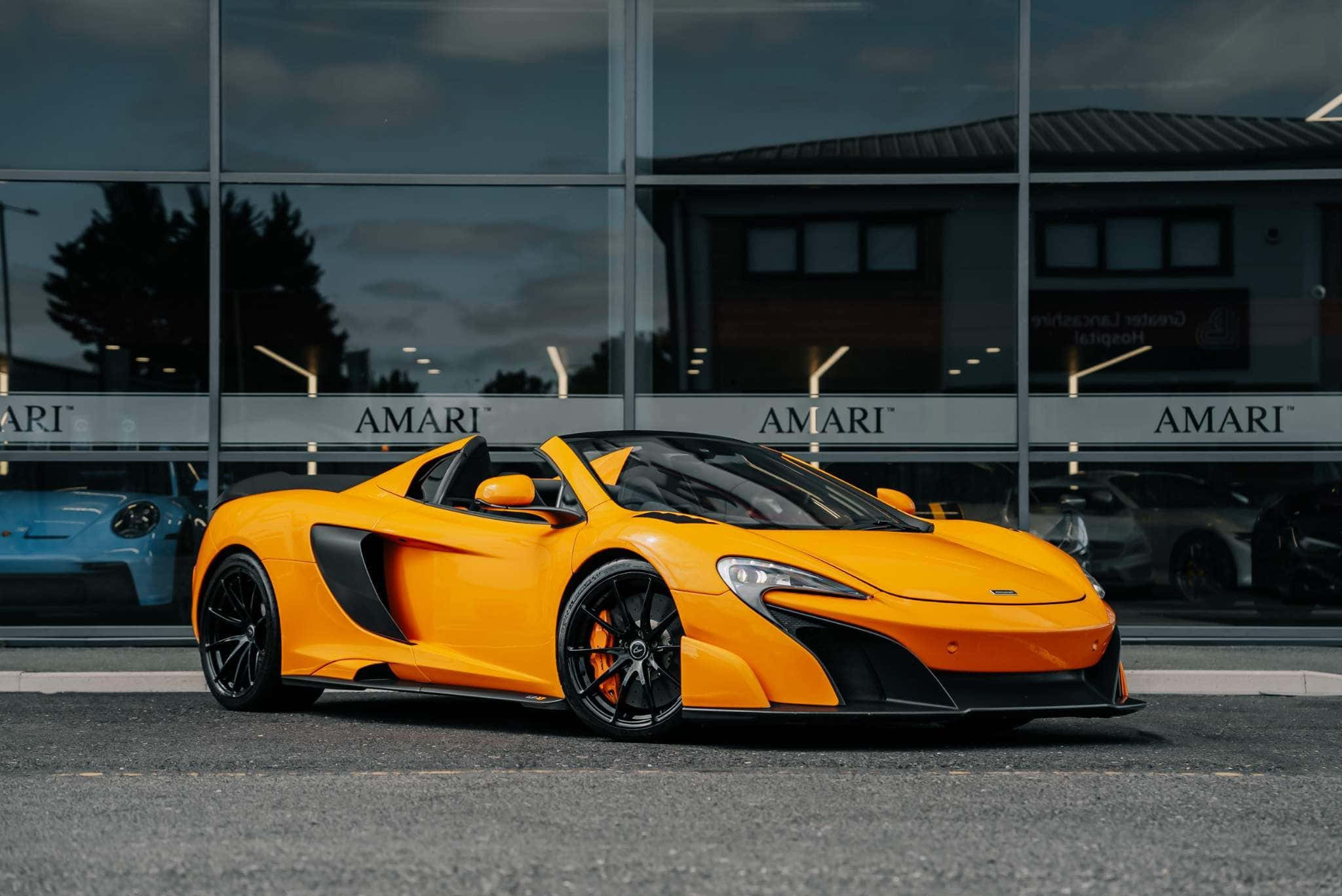 Mclaren 675lt Spider 桌布 壁紙 — 一輛鮮橙色的McLaren跑車在黃昏時停泊在玻璃幕牆的AMARI展示間外。