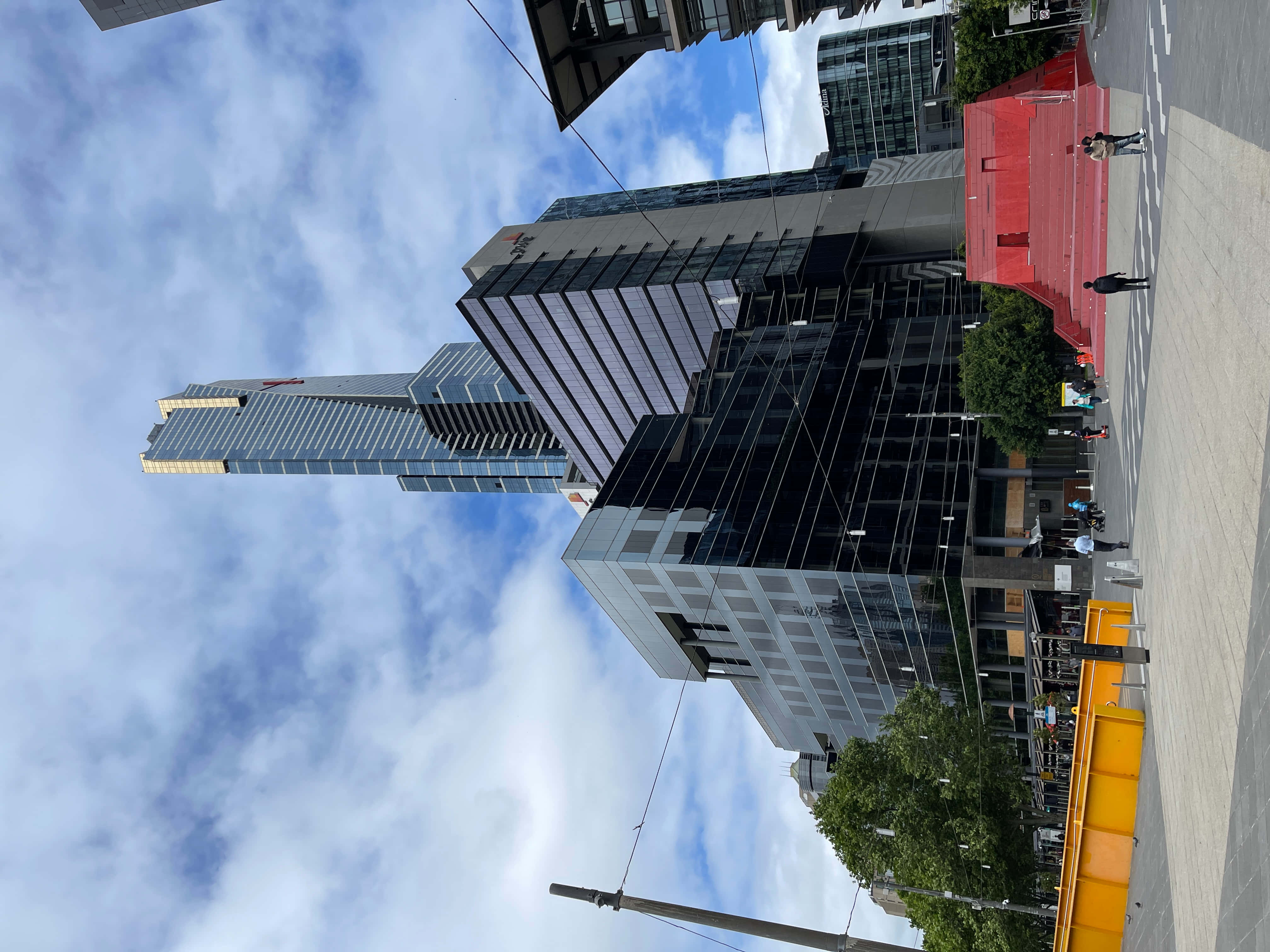 Eureka Tower 桌布 壁紙 — 現代玻璃摩天大樓在部分多雲的天空下，聳立在一個熙來攘往的城市廣場上，廣場上有行人和電車軌道。