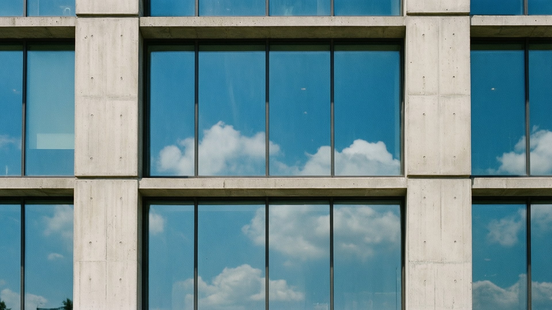 Concrete and Glass Facade 壁紙 — 現代混凝土建築外牆，配有大玻璃窗，反射出明亮的藍天和蓬鬆的白雲。