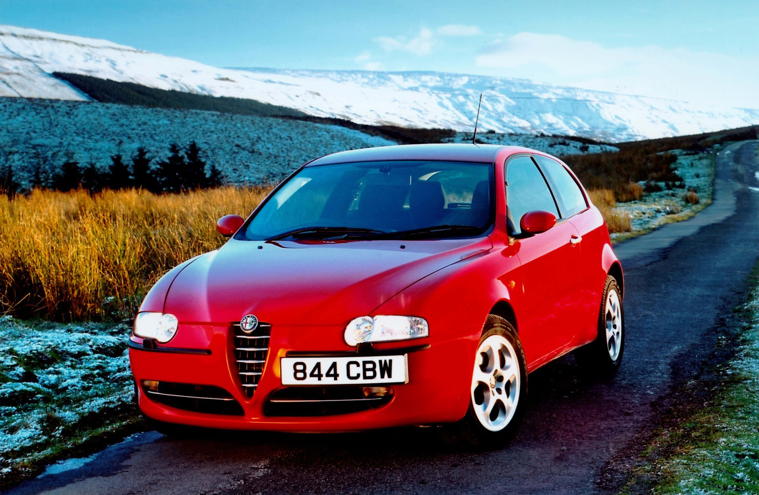 Alfa Romeo 147 桌布 壁紙 — 一輛鮮紅色的愛快羅密歐跑車停泊在蜿蜒的道路上，背景是白雪覆蓋的山脈和結霜的草地。