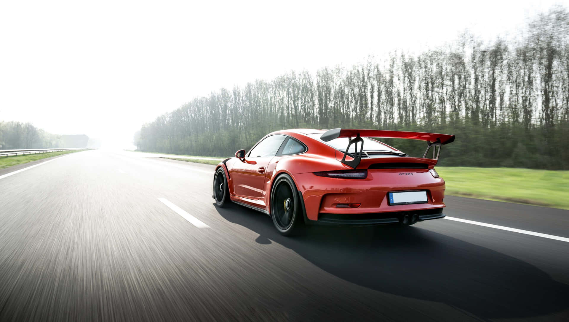 壁紙 — 一輛鮮紅色的保時捷911 GT3 RS在陽光明媚的高速公路上疾馳，帶有動態模糊效果，路邊排列著無葉的樹木。