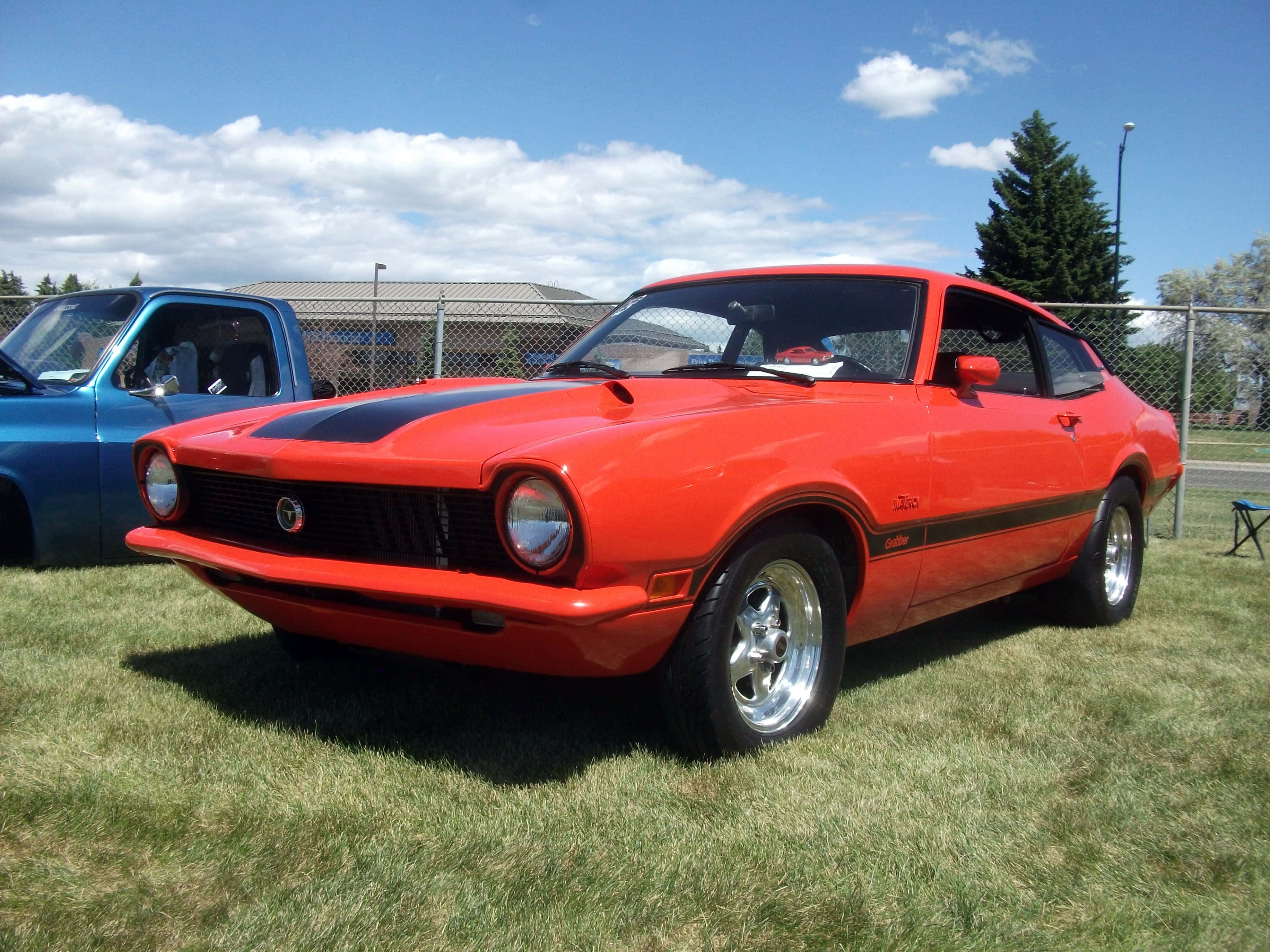 Ford Maverick 桌布 壁紙 — 一輛鮮紅色的經典肌肉車帶有黑色賽車條紋，停在草地上，天空湛藍，白雲朵朵。