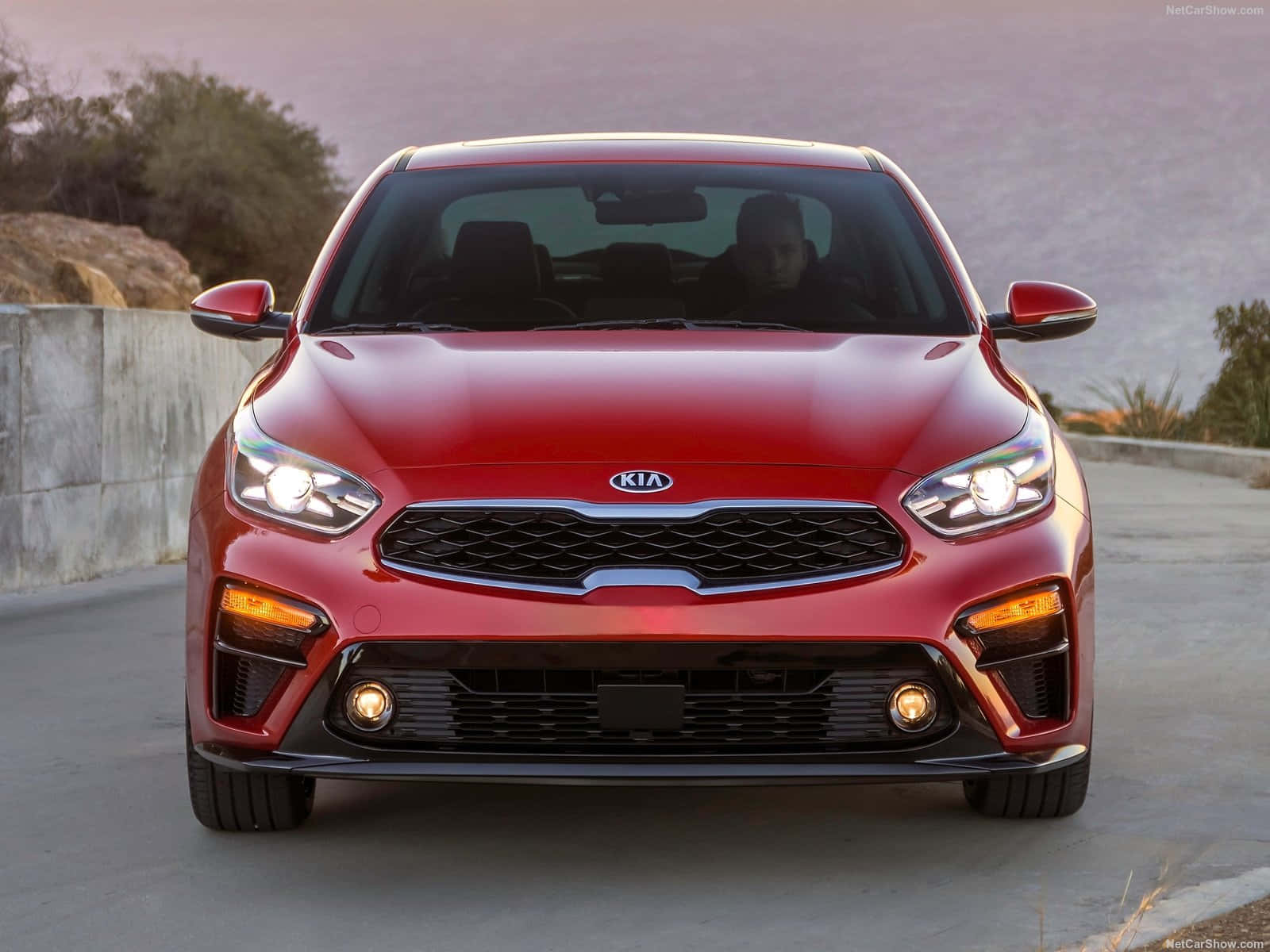 Kia Forte 桌布 壁紙 — 一輛鮮紅色的起亞汽車在日落時分的沿海公路上亮著車頭燈向前行駛。