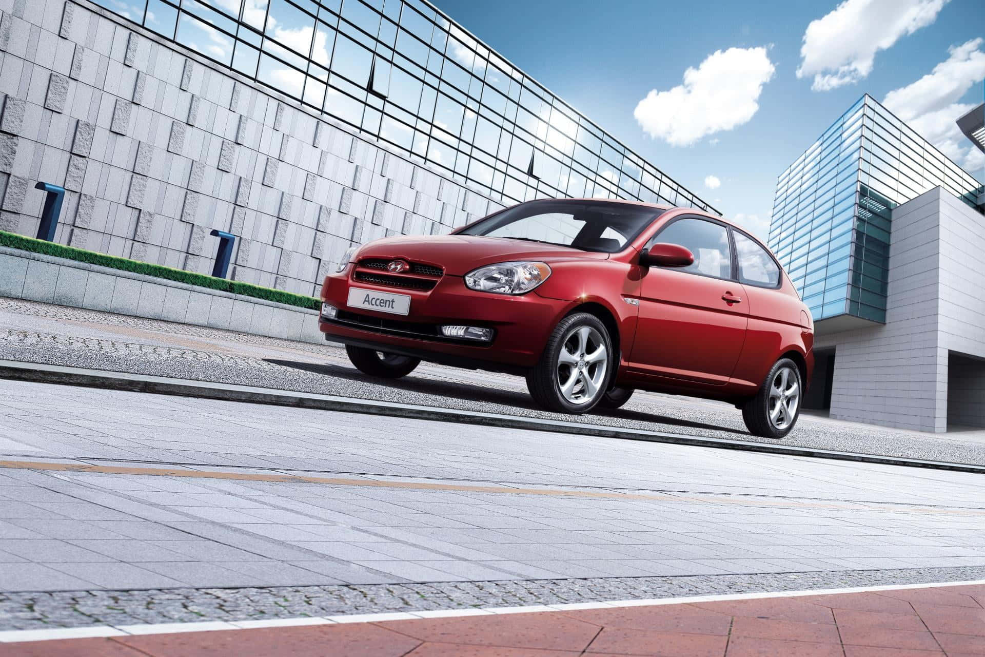 Hyundai Accent 桌布 壁紙 — 一輛鮮紅色的現代雅紳特掀背車停放在現代瓷磚廣場上，旁邊是玻璃和石頭建築，天空湛藍，白雲朵朵。