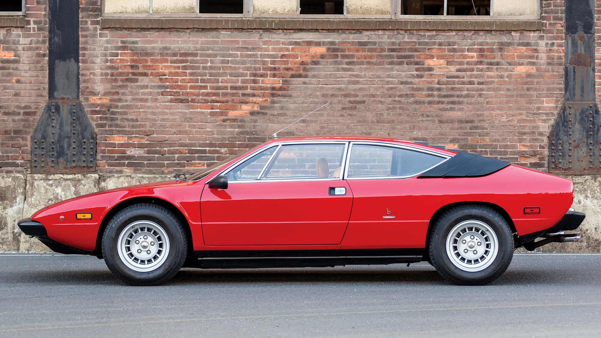 Lamborghini Urraco 桌布 壁紙 — 一輛鮮紅色的法拉利308 GTB跑車停在一棟風化的磚砌建築前，帶有工業風格的點綴。