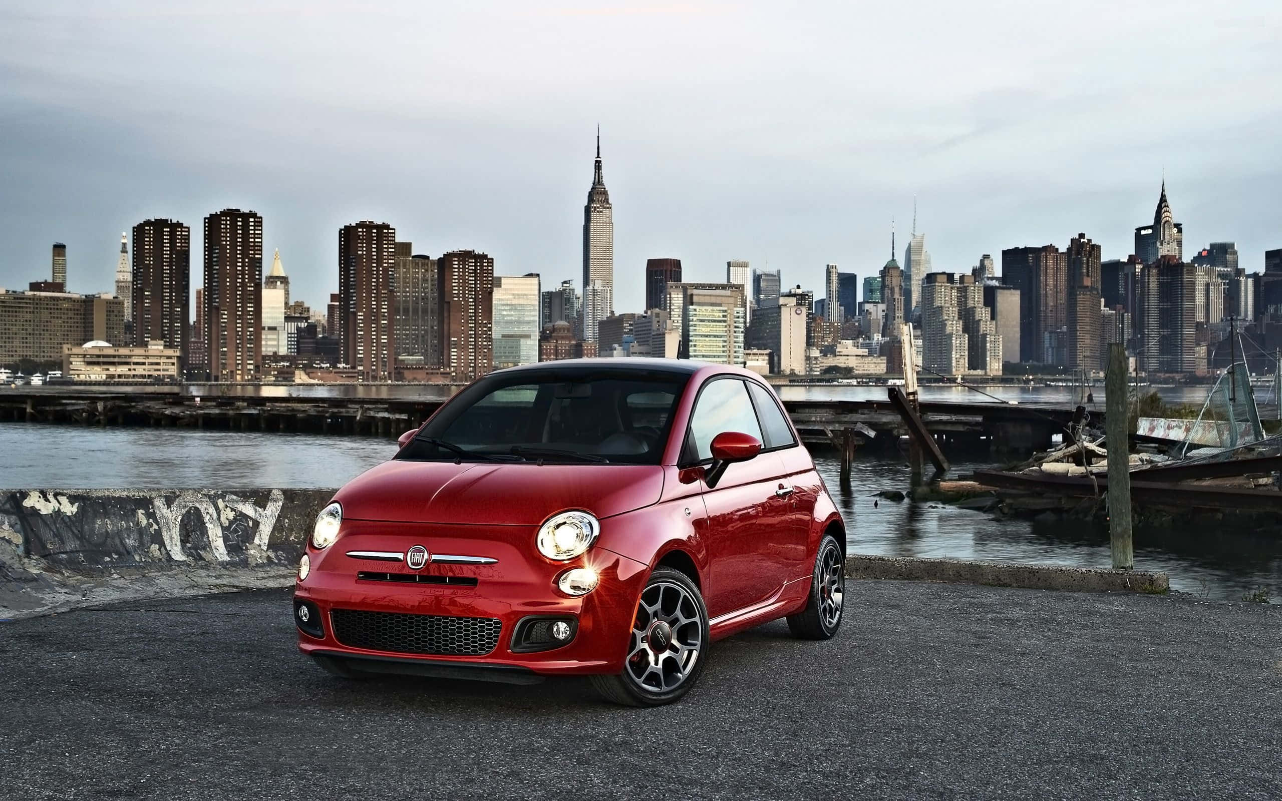 Fiat 500l 桌布 壁紙 — 一輛鮮紅色的菲亞特500停泊在海濱碼頭上，背景是陰天下的紐約市標誌性天際線。