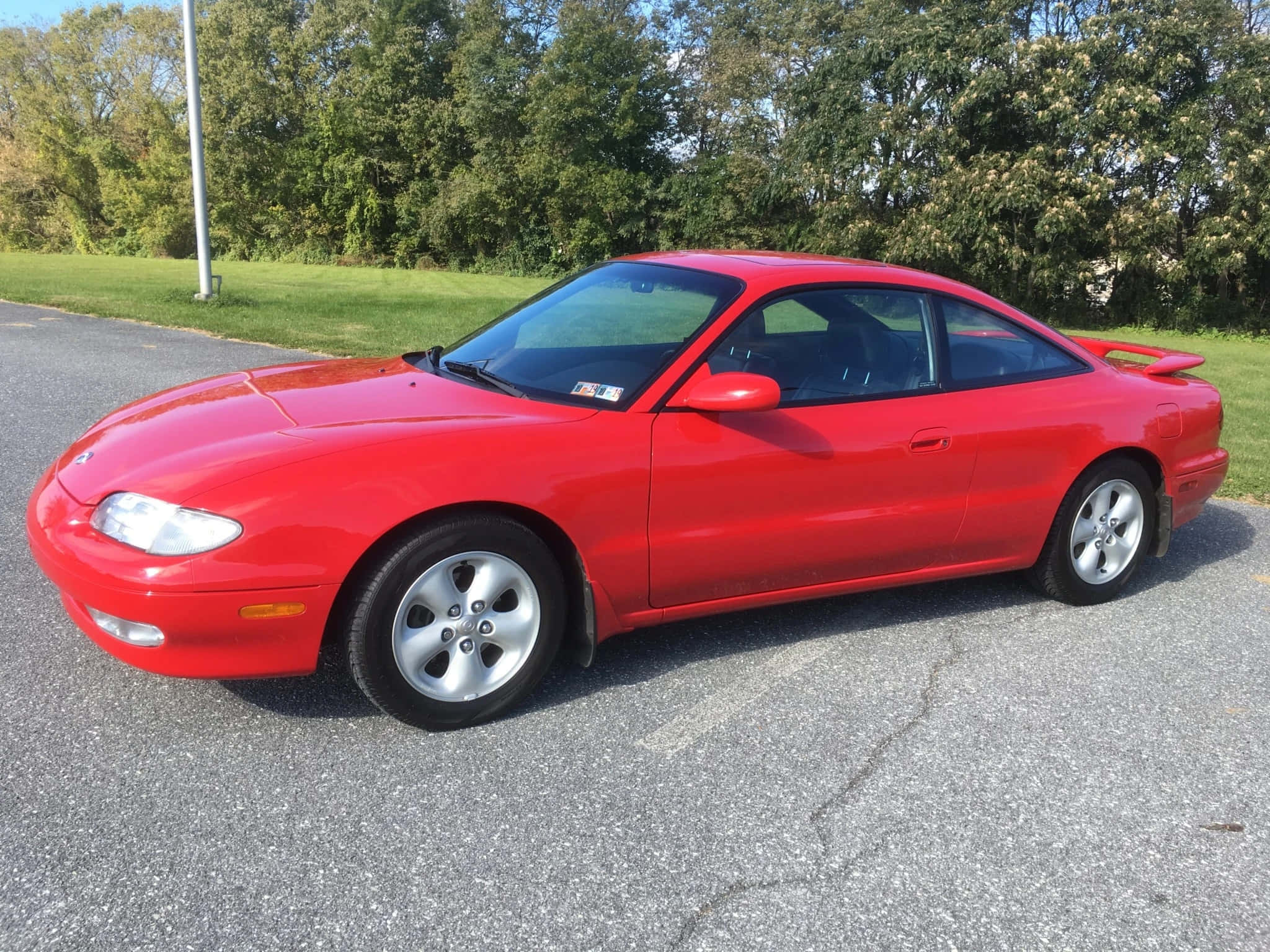 Mazda Mx-6 桌布 壁紙 — 一輛鮮紅色的馬自達MX-5跑車停在裂開的柏油路上，背景有綠樹和草地，在明亮的日光下。