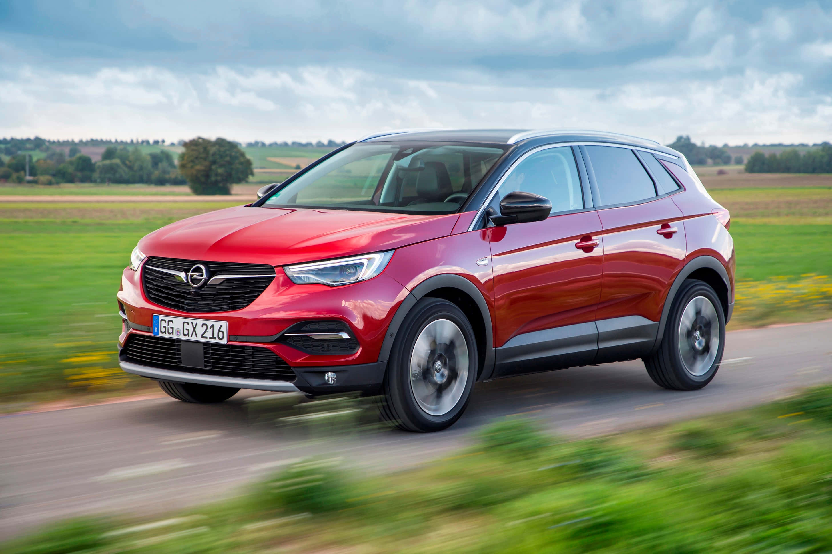 Opel Grandland X 桌布 壁紙 — 一輛鮮紅色的Opel SUV在鄉村道路上疾駛，背景是模糊的綠色田野和多雲的天空。