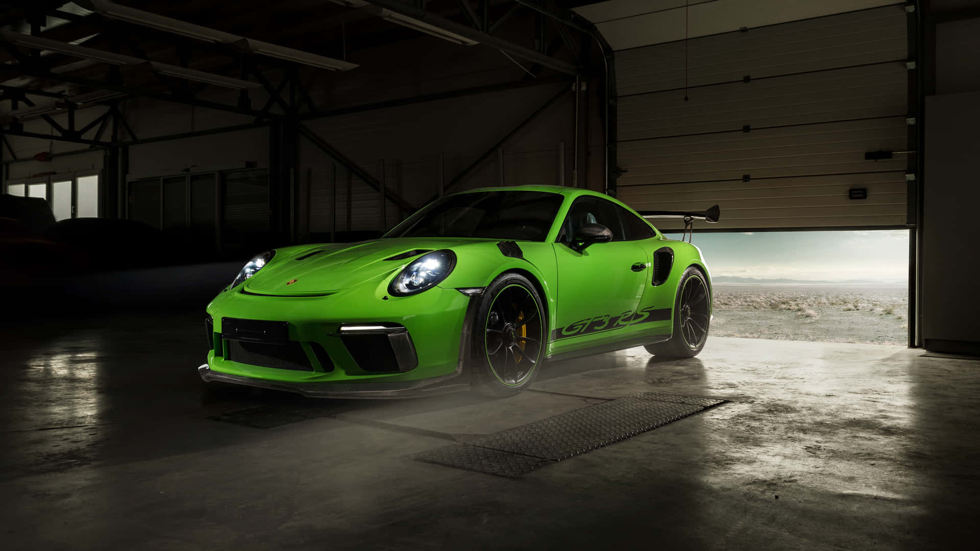  壁紙 — 一輛鮮綠色保時捷911 GT3 RS在寬敞的工業車庫內，在工作室燈光下閃閃發光，車庫外可看到沙漠景色。