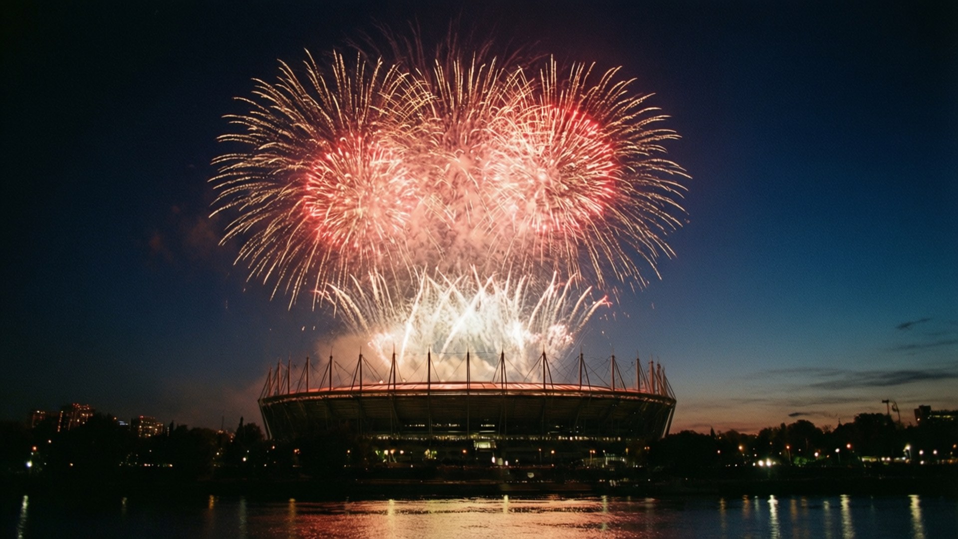 Fireworks Celebration 壁紙 — 鮮豔的紅色和白色煙火在黃昏時分在平靜的水面倒映的現代體育場上空爆炸。