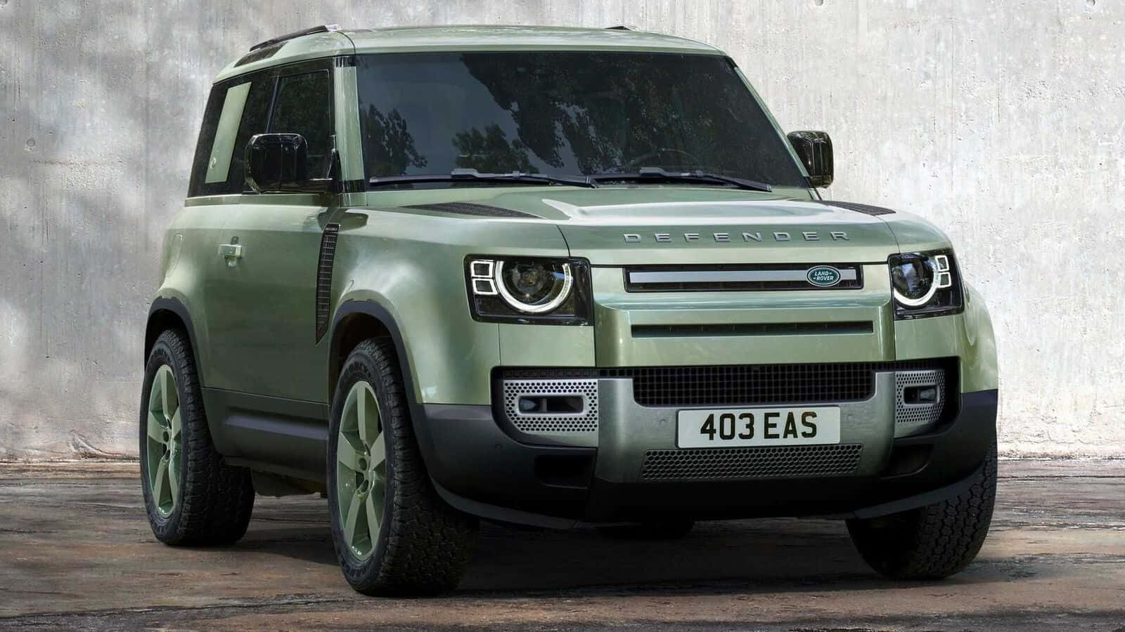 Land Rover Defender 桌布 壁紙 — 一輛消光橄欖綠的Land Rover Defender在明亮的日光下，大膽地停在一面風化的混凝土牆前。