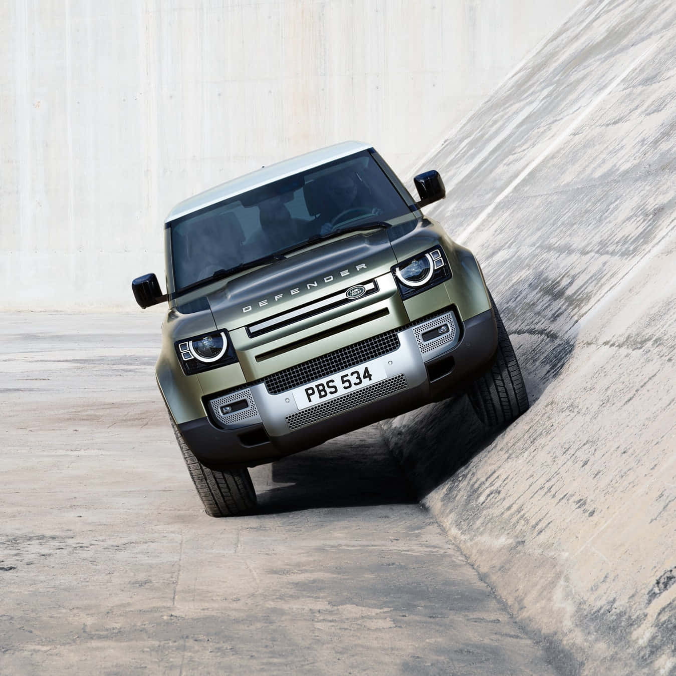Land Rover Defender 桌布 壁紙 — 一輛消光橄欖綠色的 Land Rover Defender 在簡樸的工業環境中急劇地傾斜在混凝土斜坡上。