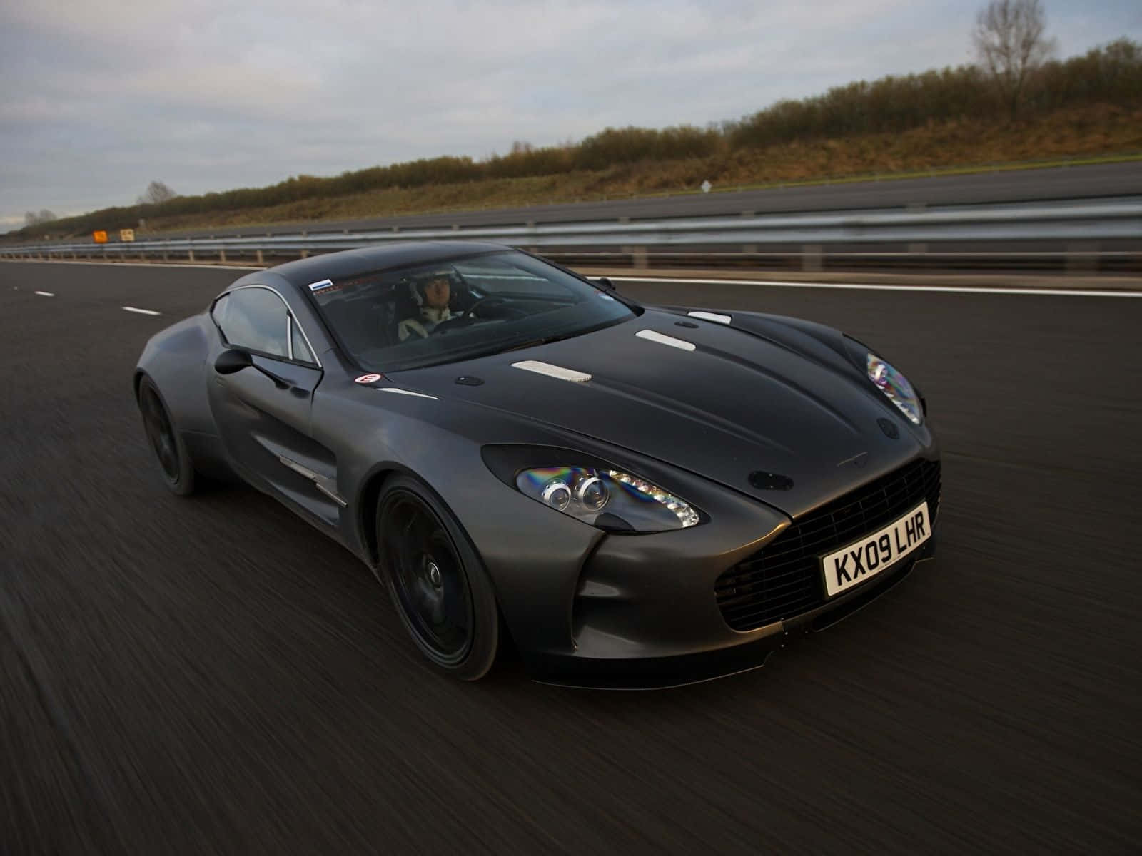 Aston Martin One-77 桌布 壁紙 — 一輛消光黑色的Aston Martin V12 Vantage在高速公路上奔馳，帶有動態模糊和陰天。