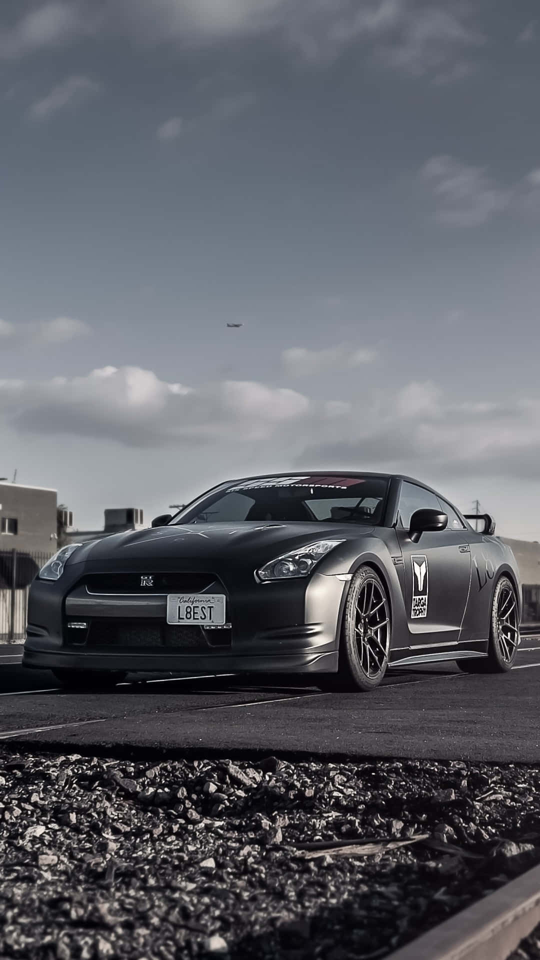 消光黑色日產GT-R停在瀝青上，前景有碎石，天空多雲。 — 吉他 iPhone 壁紙