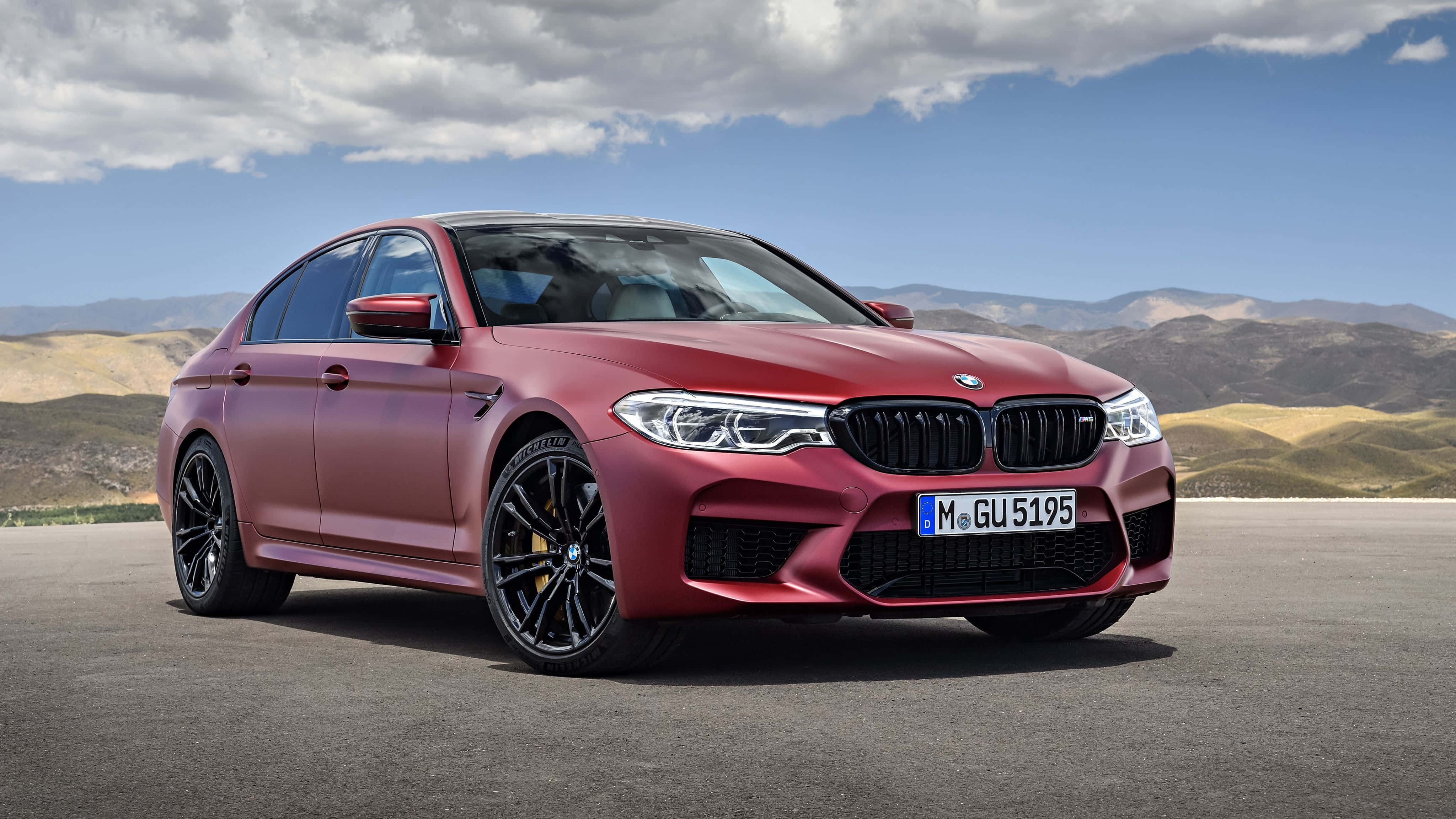 Bmw M5 桌布 壁紙 — 一輛消光紅色BMW M5轎車停在柏油路上，背景有山脈和多雲的天空。