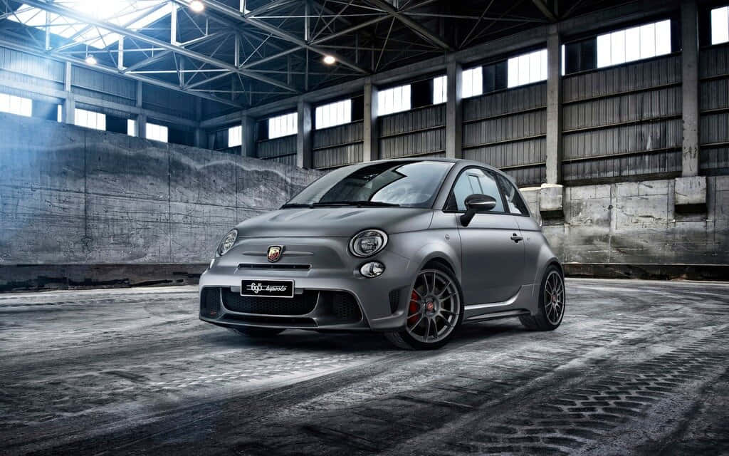 Fiat 500 桌布 壁紙 — 一輛消光灰色的Fiat 500 Abarth跑車，帶有紅色制動卡鉗，停在一個粗糙的工業倉庫中，天花板很高，地板是混凝土的。
