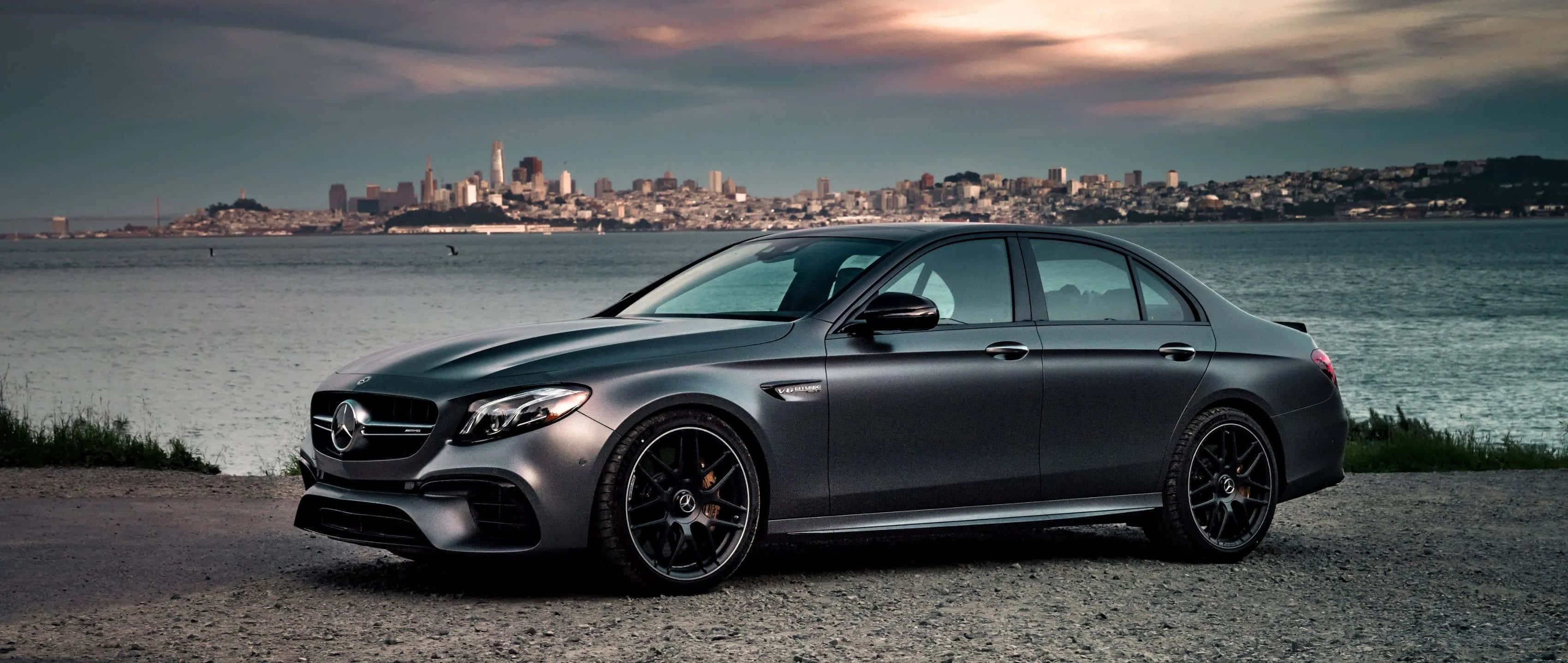 Coupe 桌布 壁紙 — 一輛消光灰色的梅賽德斯-AMG E63轎車停在礫石岸邊，水對岸的城市天際線在陰沉的暮色天空下。