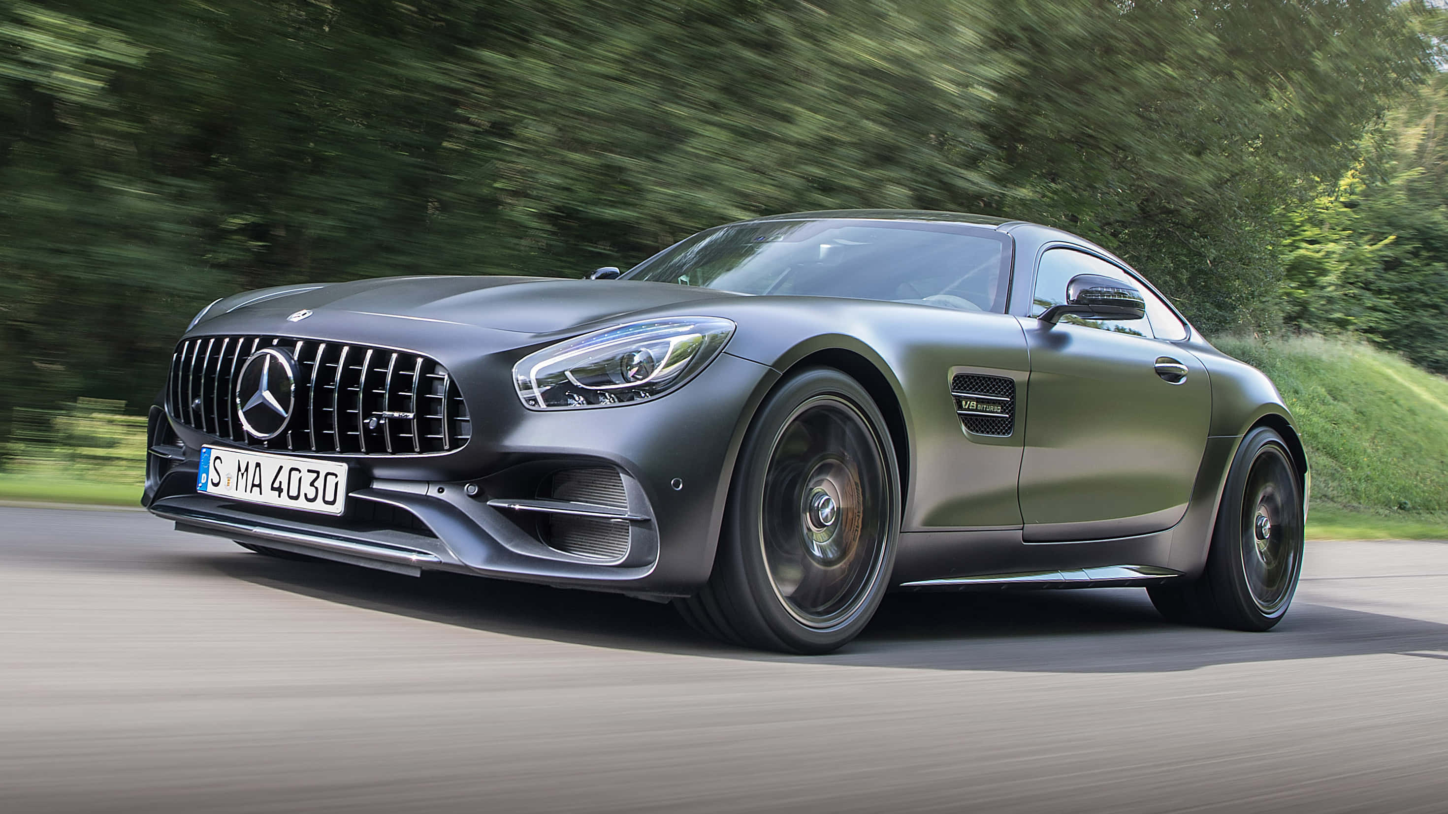 Mercedes Benz Amg Gt 桌布 壁紙 — 一輛消光灰色的 Mercedes-AMG GT R 跑車在模糊的綠色森林背景的道路上疾馳。