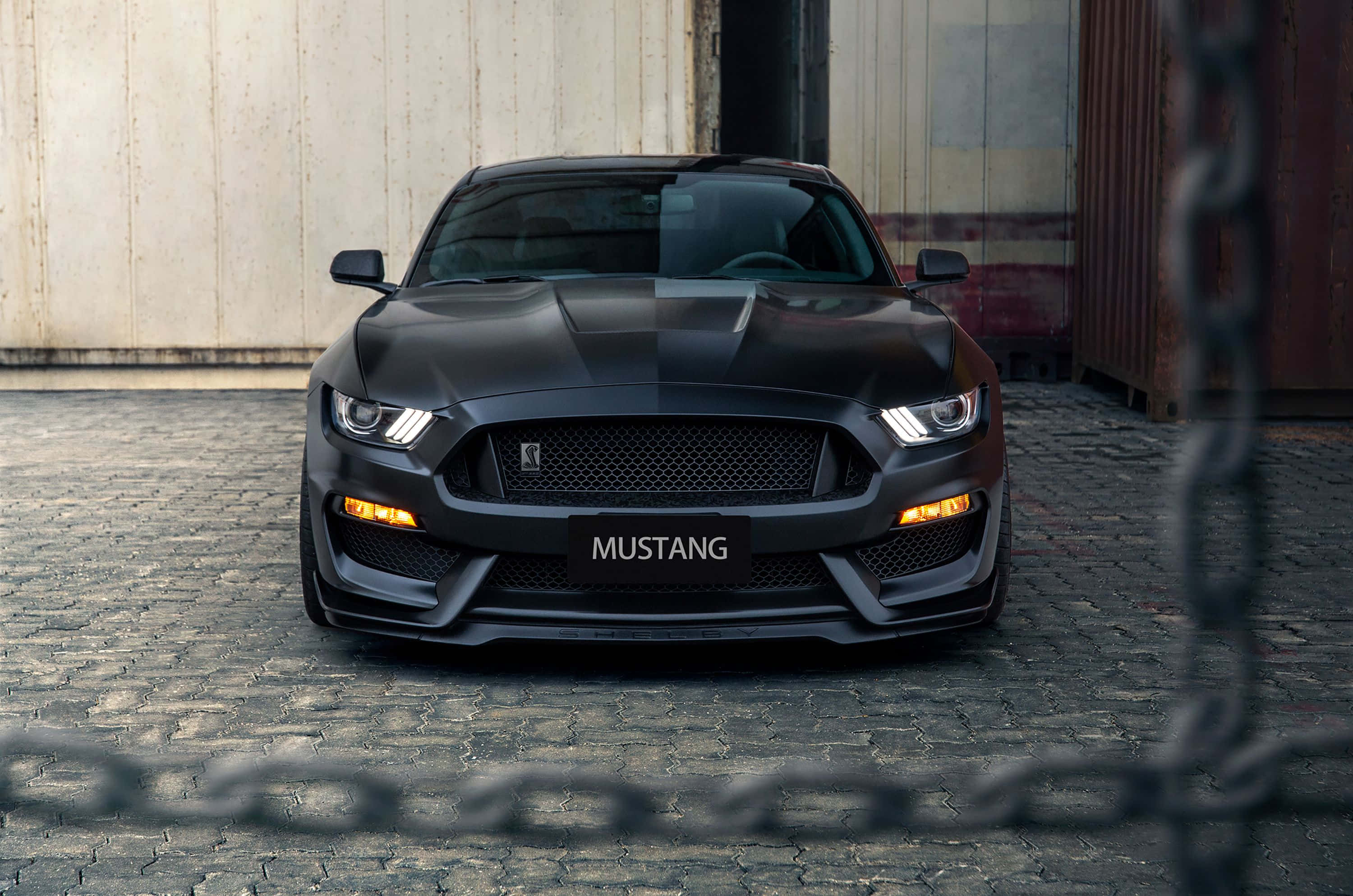 Ford Mustang Shelby Gt350 桌布 壁紙 — 一輛時尚的啞光黑色福特野馬跑車，車頭燈發光，停在工業貨櫃前的鵝卵石街道上。