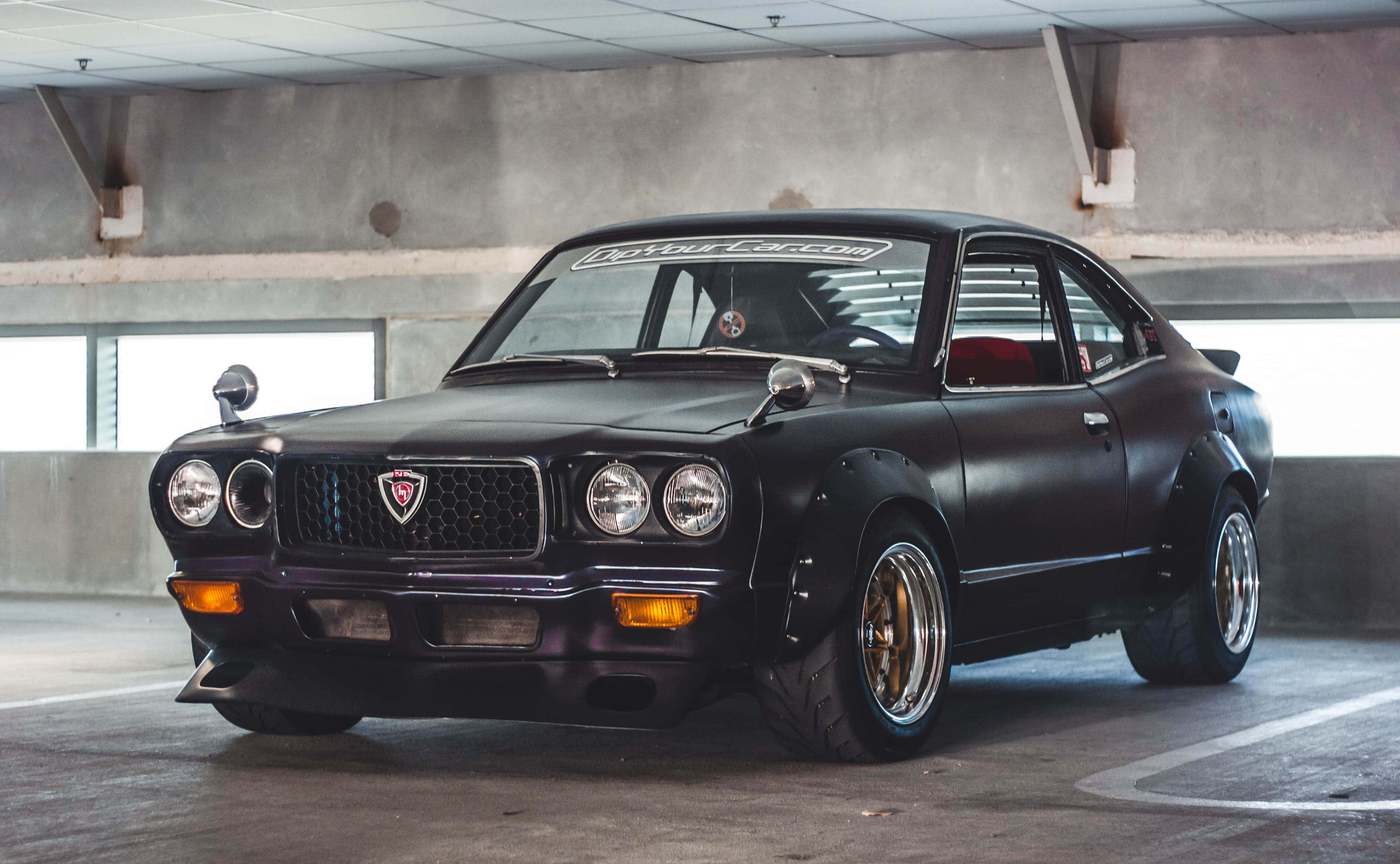 Mazda Rx-3 桌布 壁紙 — 一輛啞光黑色經典日本雙門跑車配金色輪圈停放在混凝土停車場。