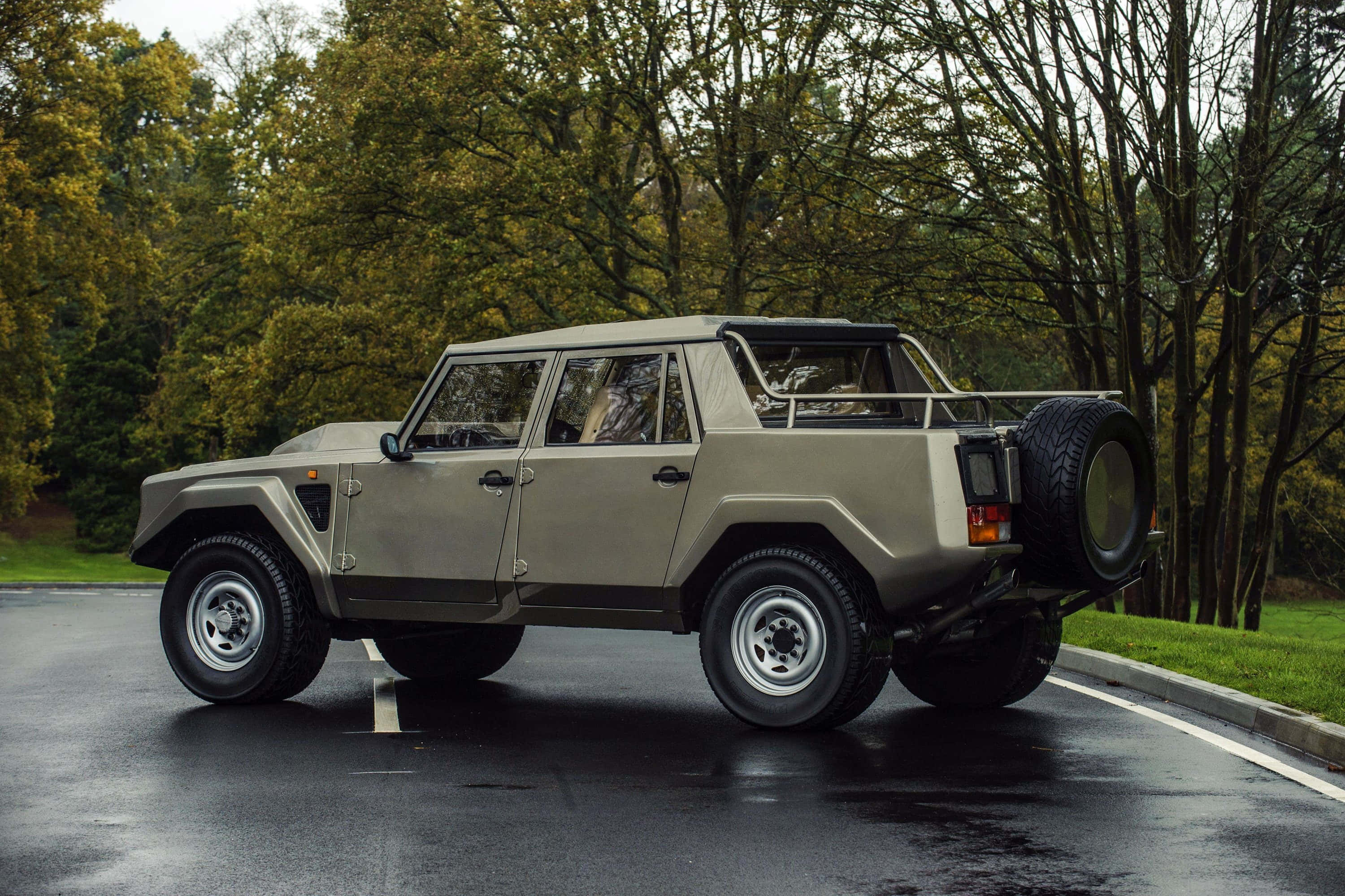 Lamborghini Lm002 桌布 壁紙 — 一輛啞光米色藍寶堅尼LM002 SUV停泊在潮濕的瀝青路上，旁邊是帶有秋樹的草地。