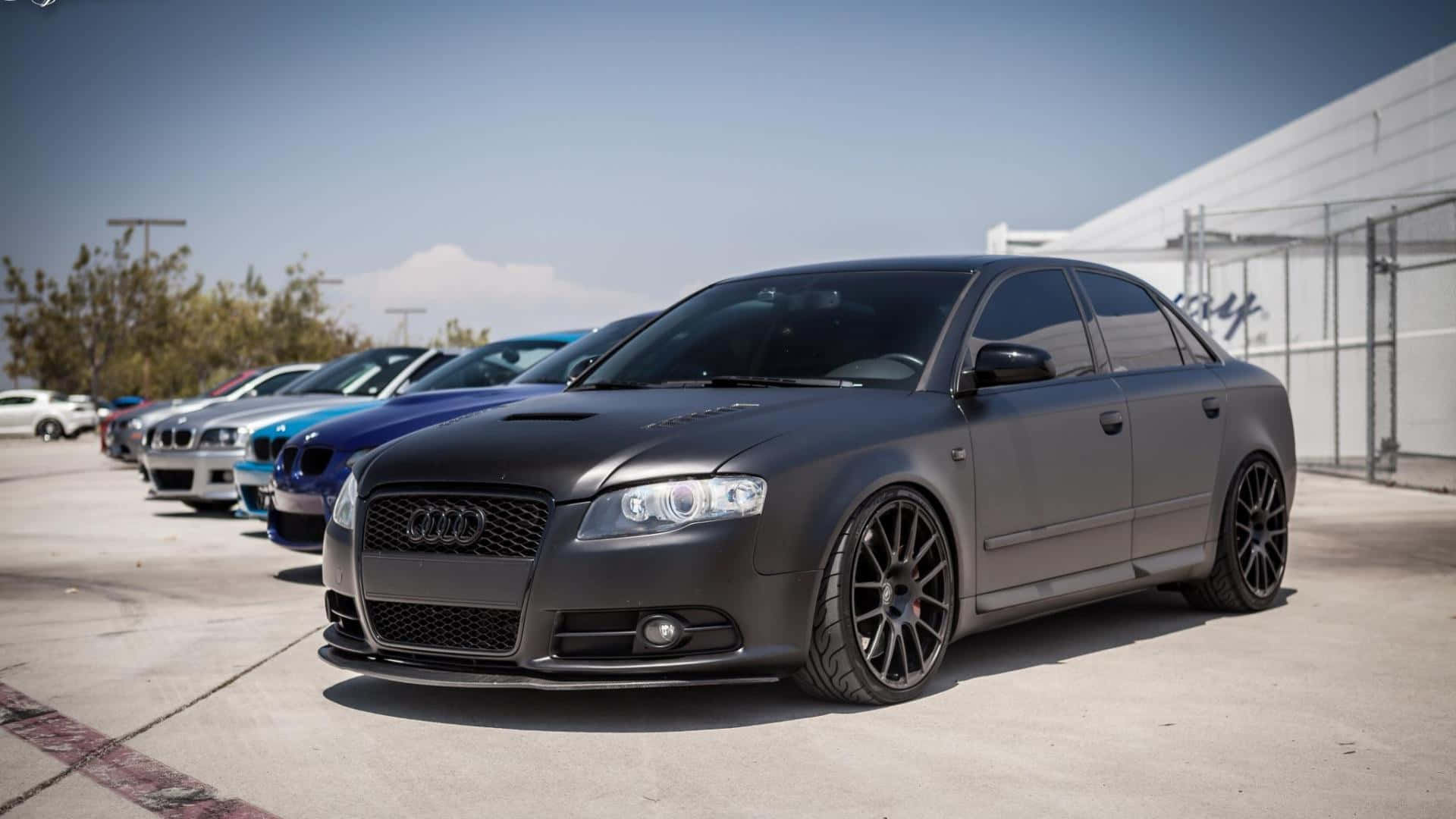Audi Rs4 桌布 壁紙 — 一輛啞黑色奧迪轎車配備訂製輪圈，在明亮日光下排成一列停泊。