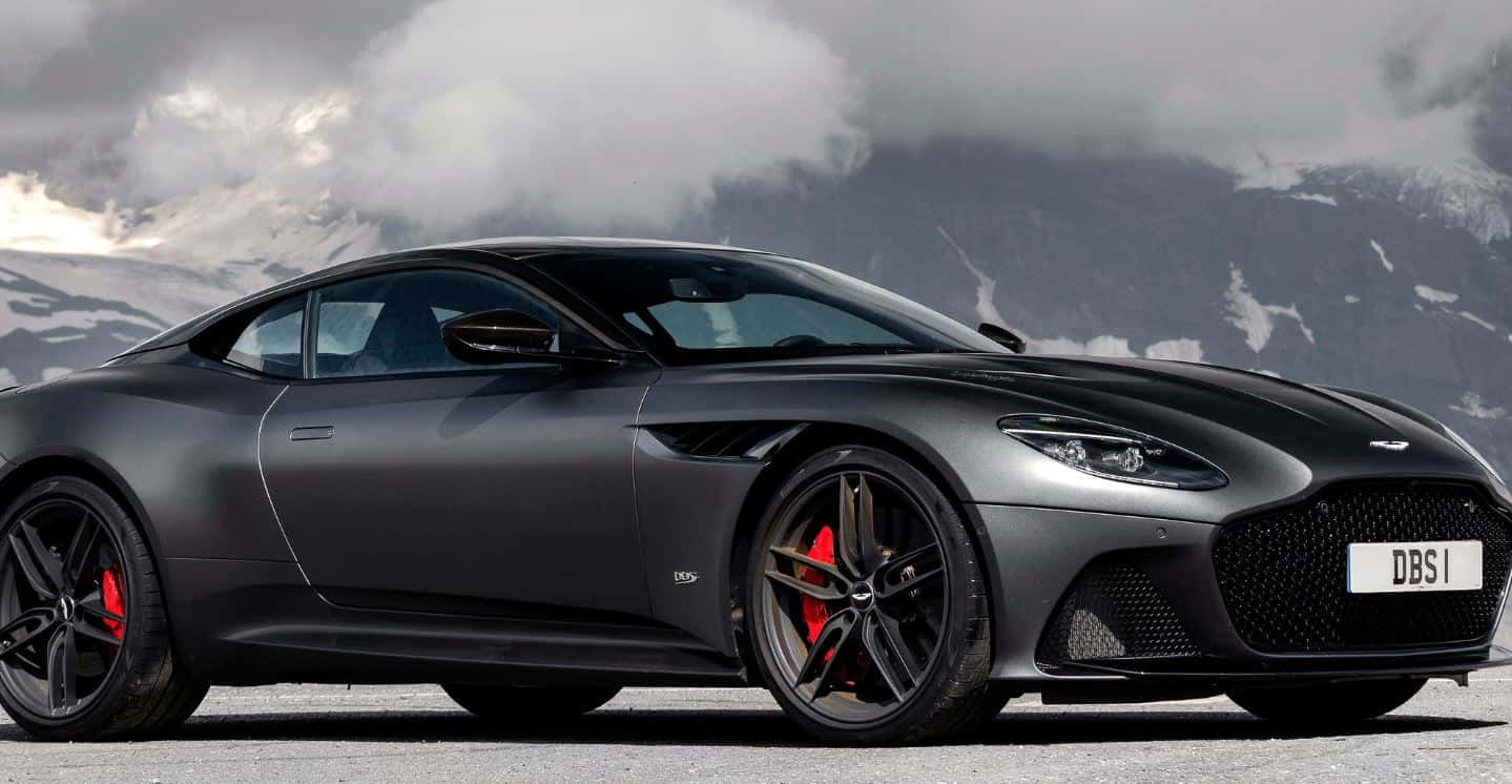 Aston Martin Dbs Superleggera 桌布 壁紙 — 一架時尚的啞黑色Aston Martin DBS1超級跑車，配有紅色制動卡鉗，停在積雪的山路上，天空有戲劇性的雲層。