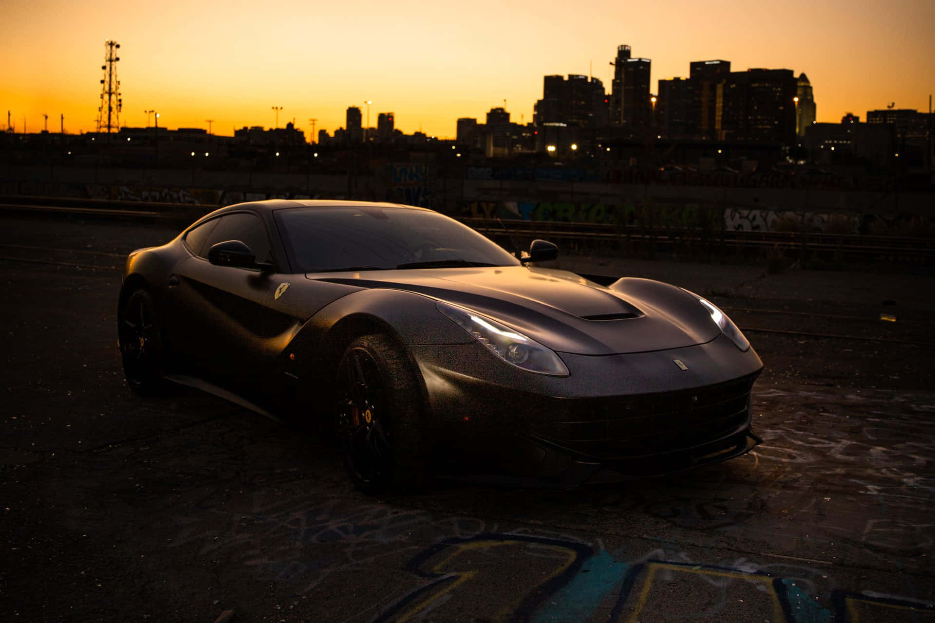 Ferrari F12 Berlinetta 桌布 壁紙 — 一輛啞黑色法拉利F12 Berlinetta停泊在日落時分被塗鴉覆蓋的城市空地，背景是城市天際線。