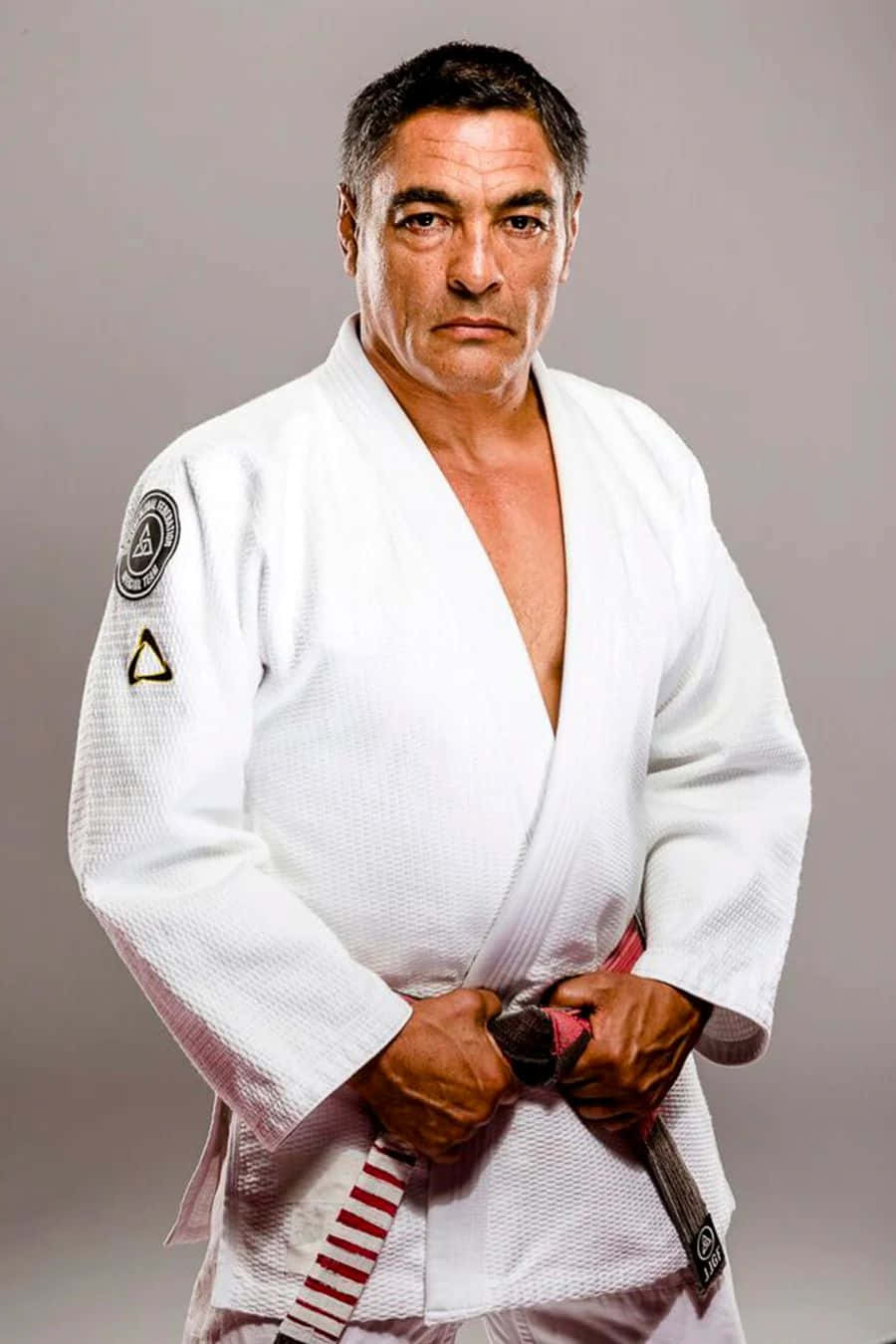 Rickson Gracie 桌布 壁紙 — 一位嚴肅的中年男子穿著白色柔道服，繫著紅帶，自信地站在純灰色背景前。