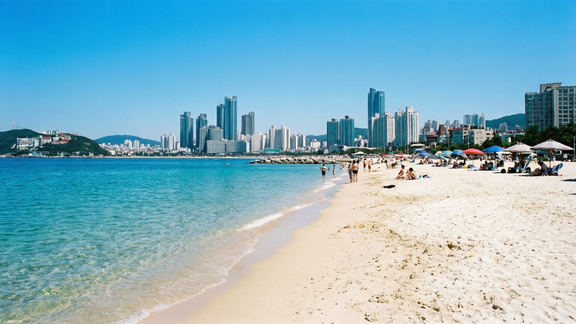 Busan Haeundae Summer 壁紙 — 陽光明媚的海灘，碧綠海水，輕柔海浪，沙灘，背景是現代城市天際線。