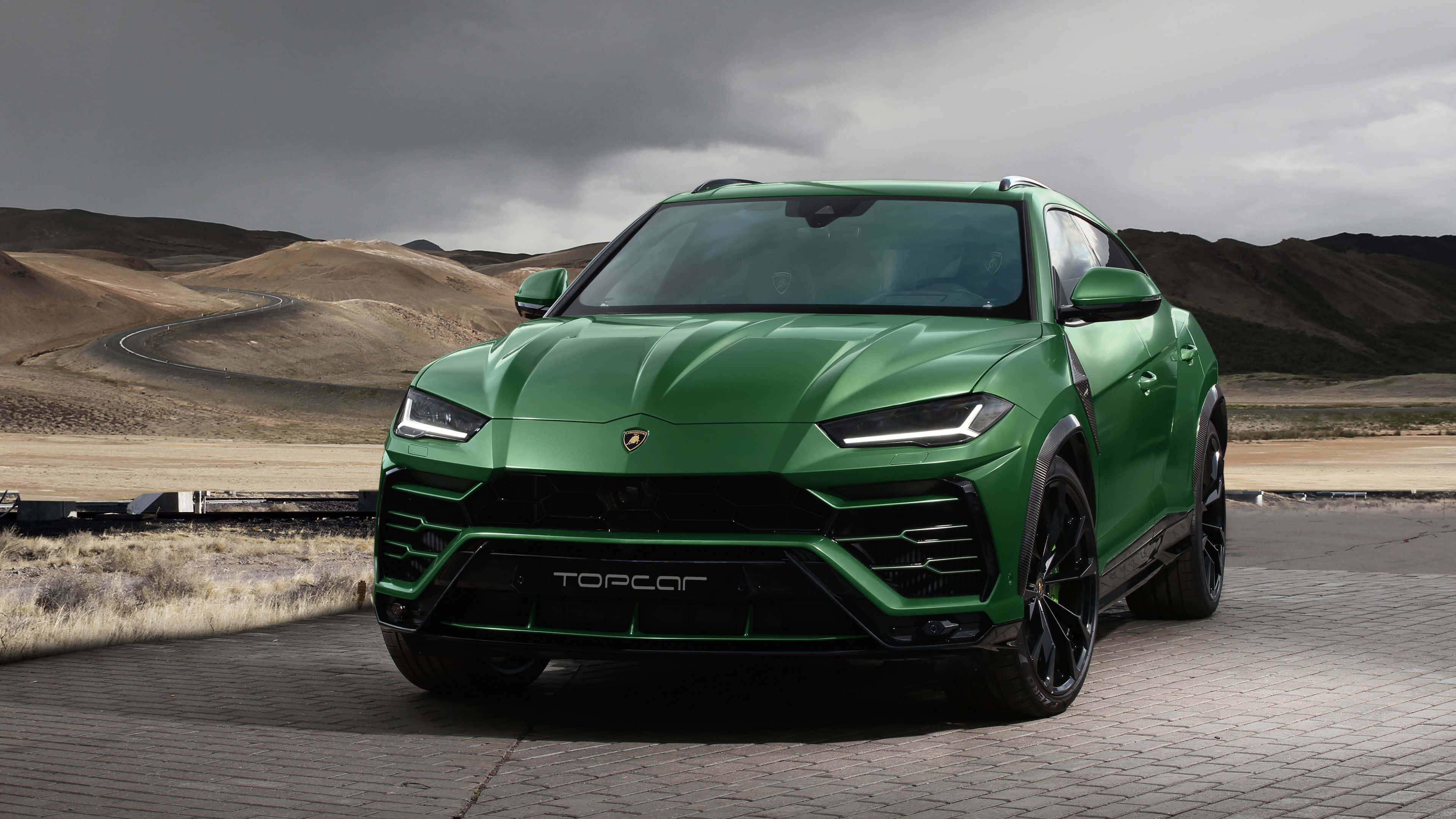 Lamborghini Urus 桌布 壁紙 — 一輛充滿活力的綠色藍寶堅尼 Urus Performante 停在鋪砌的觀景點上，背景是戲劇性的多雲天空和荒蕪的山丘。